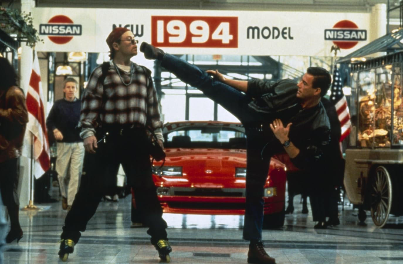 Zwei Männer, einer tritt dem anderen ins Gesicht. Im Hintergrund steht ein rotes Auto mit einem Schild "1994 Model".