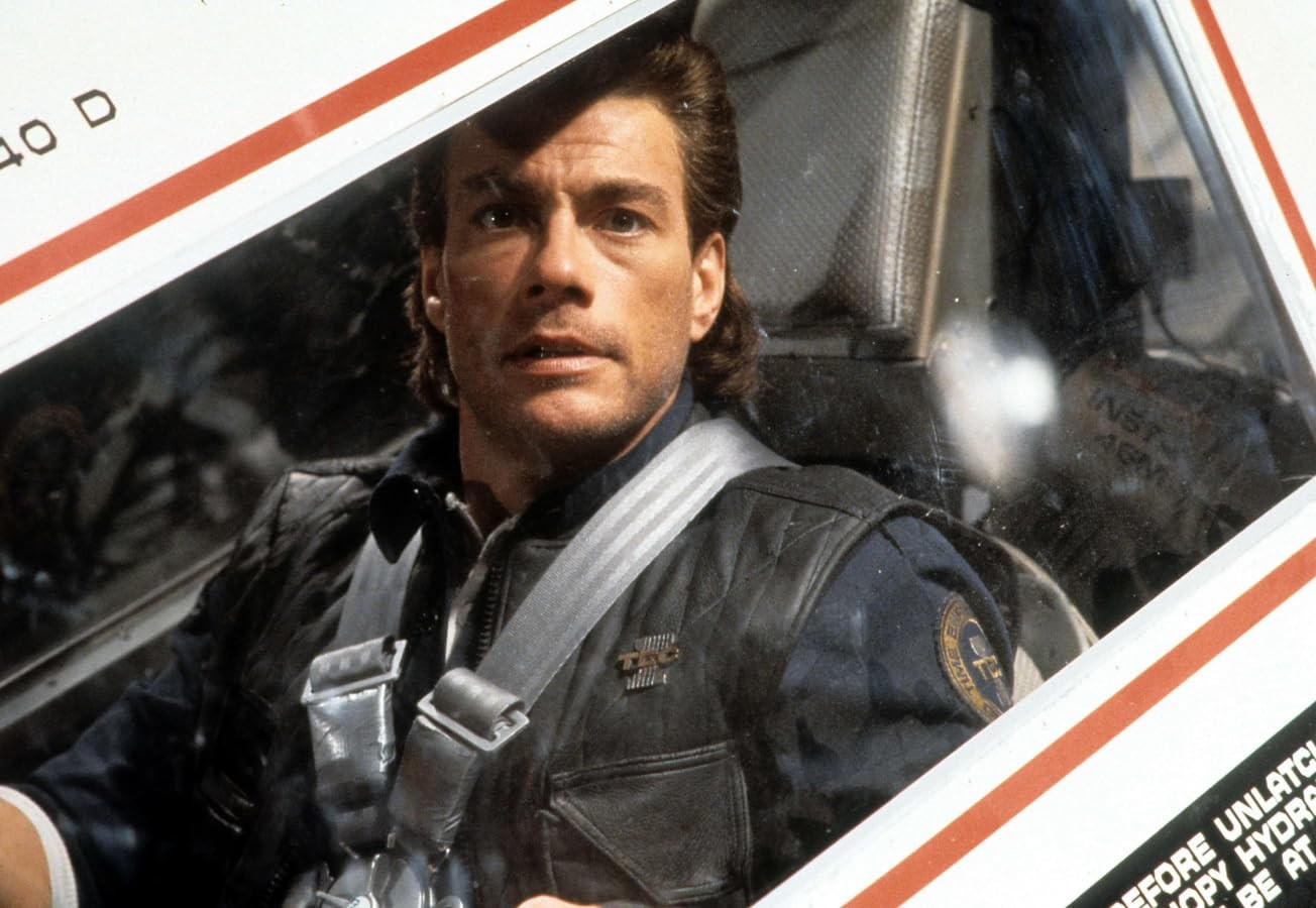 Jean-Claude Van Damme in einem Cockpit, trägt eine schwarze Lederjacke und einen Sicherheitsgurt, blickt ernst nach vorne.