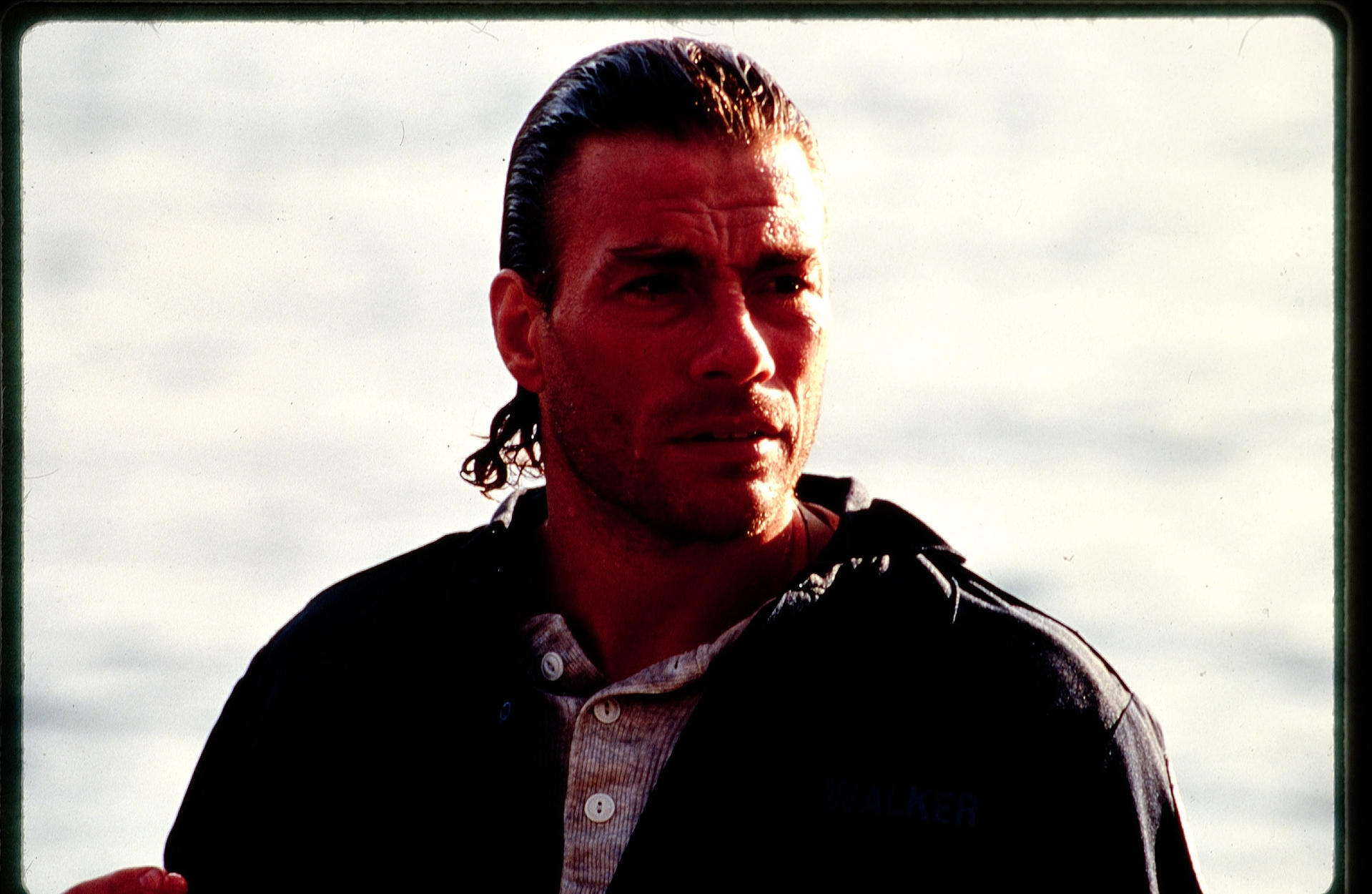 Ein Mann mit zurückgegeltem Haar und Jacke blickt zur Seite. Der Hintergrund ist ein hellfarbener Himmel.
