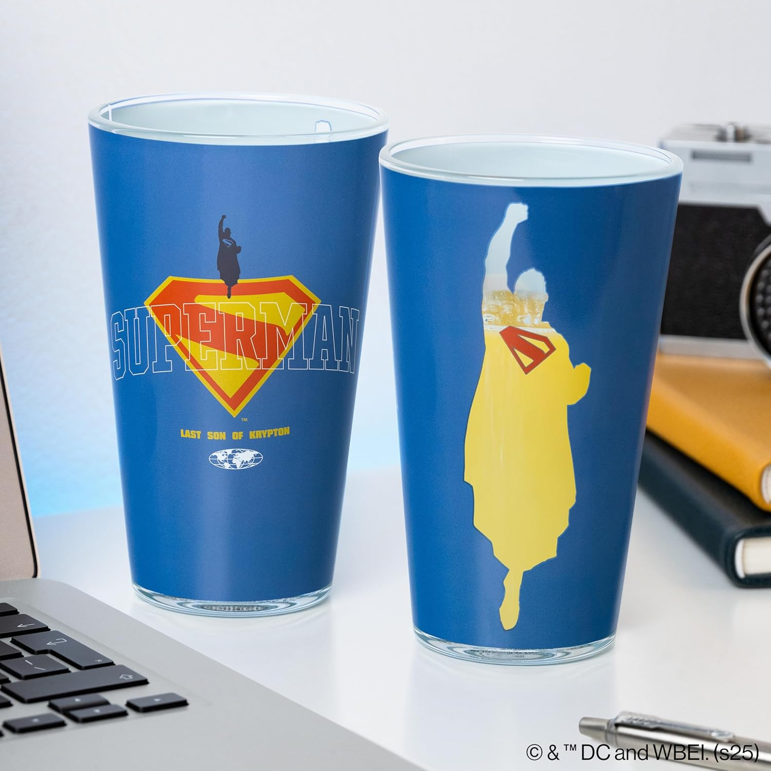 Két kék Superman pohár. Az egyiken Superman logó, a másikon sárga sziluett. Egy laptop és egy kamera a fehér felületen.