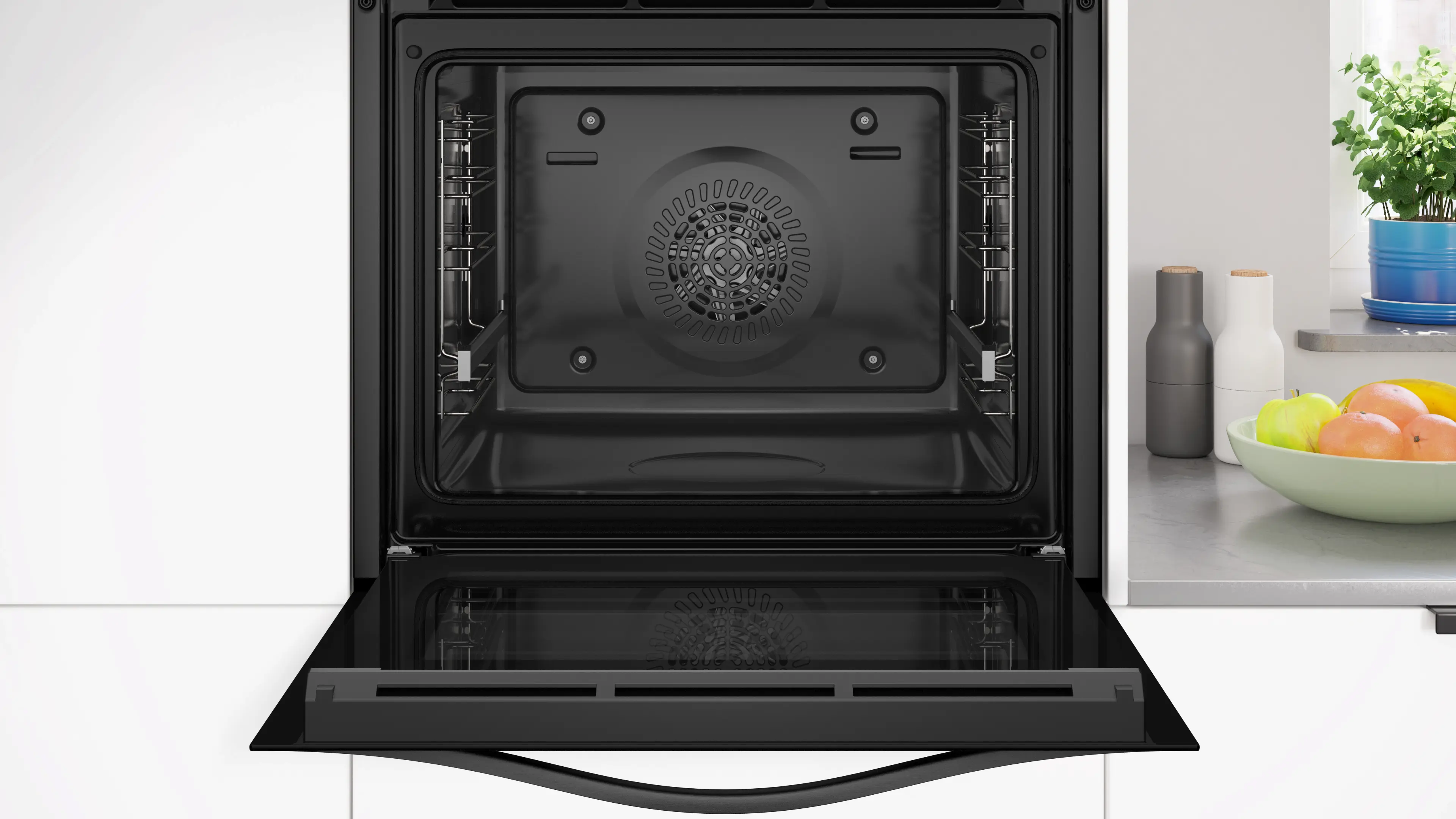 Horno abierto con puerta negra. El interior es oscuro con un ventilador. Frutas, plantas y botellas están en la encimera a la derecha.