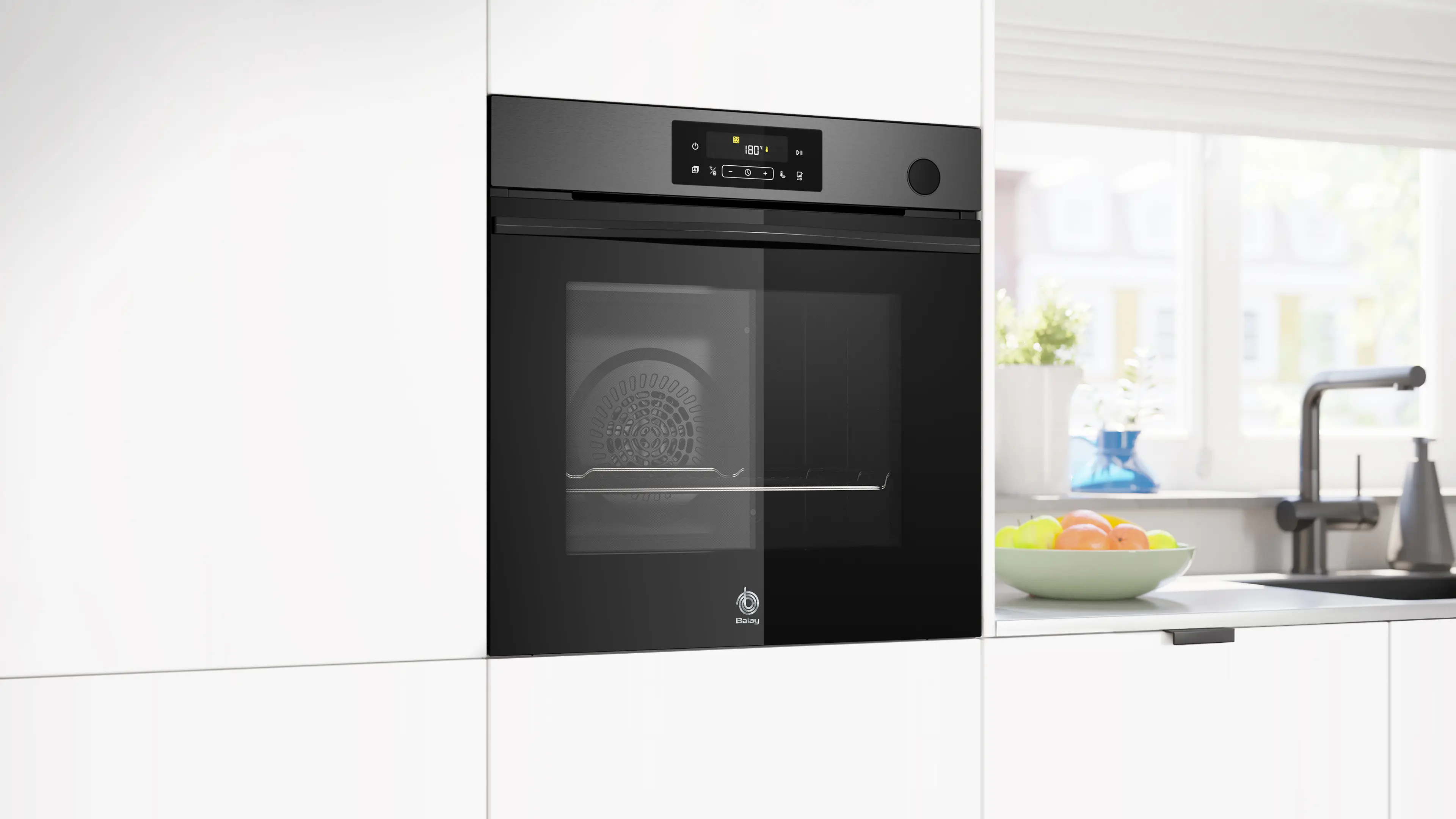 Un horno negro empotrado es el punto focal en una cocina blanca. Hay fruta en un cuenco sobre la encimera. Hay una ventana en el fondo.