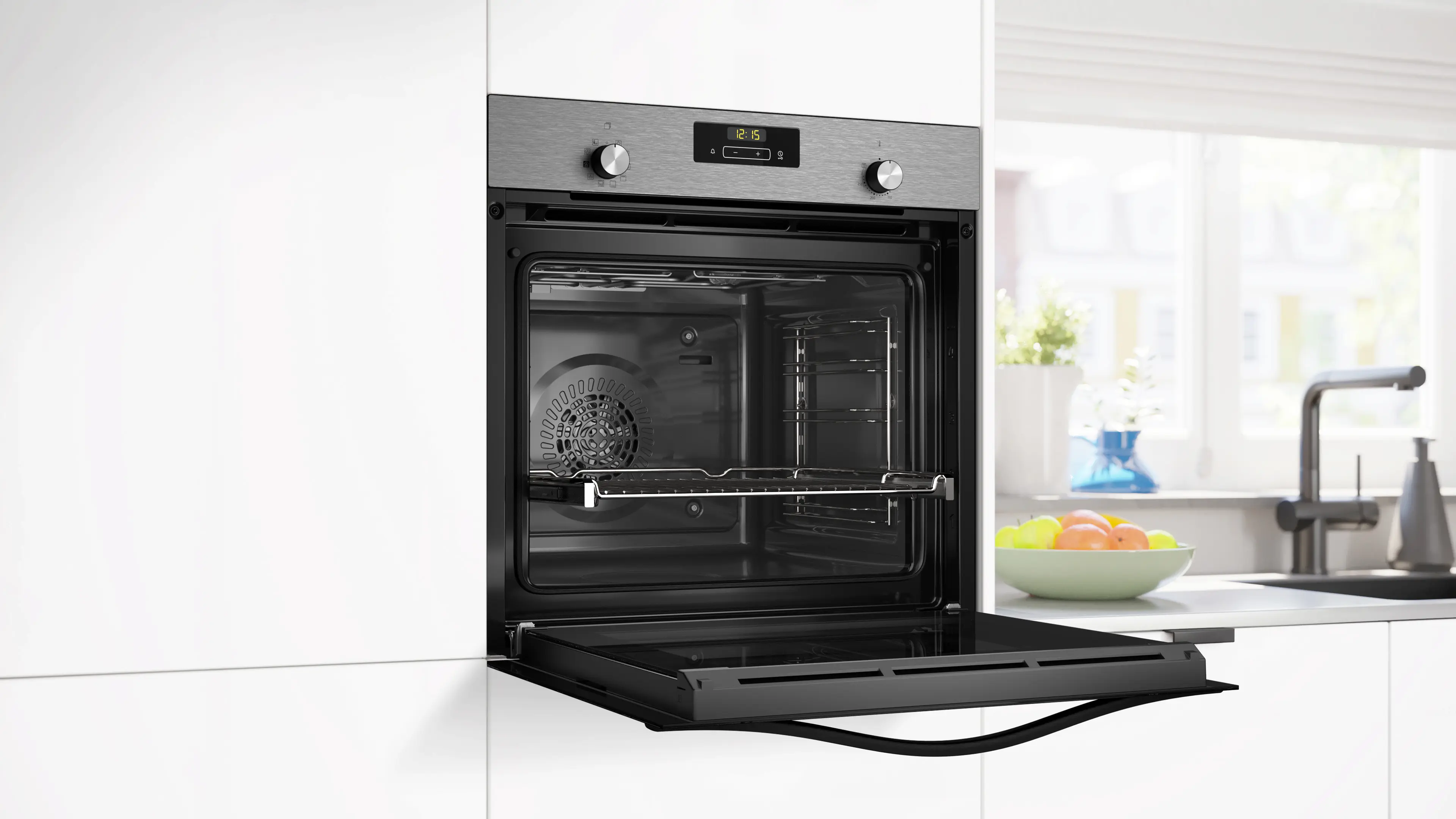 Un horno con la puerta abierta está en una cocina. El horno es de acero inoxidable con reloj digital. Un bol de fruta está en la encimera.