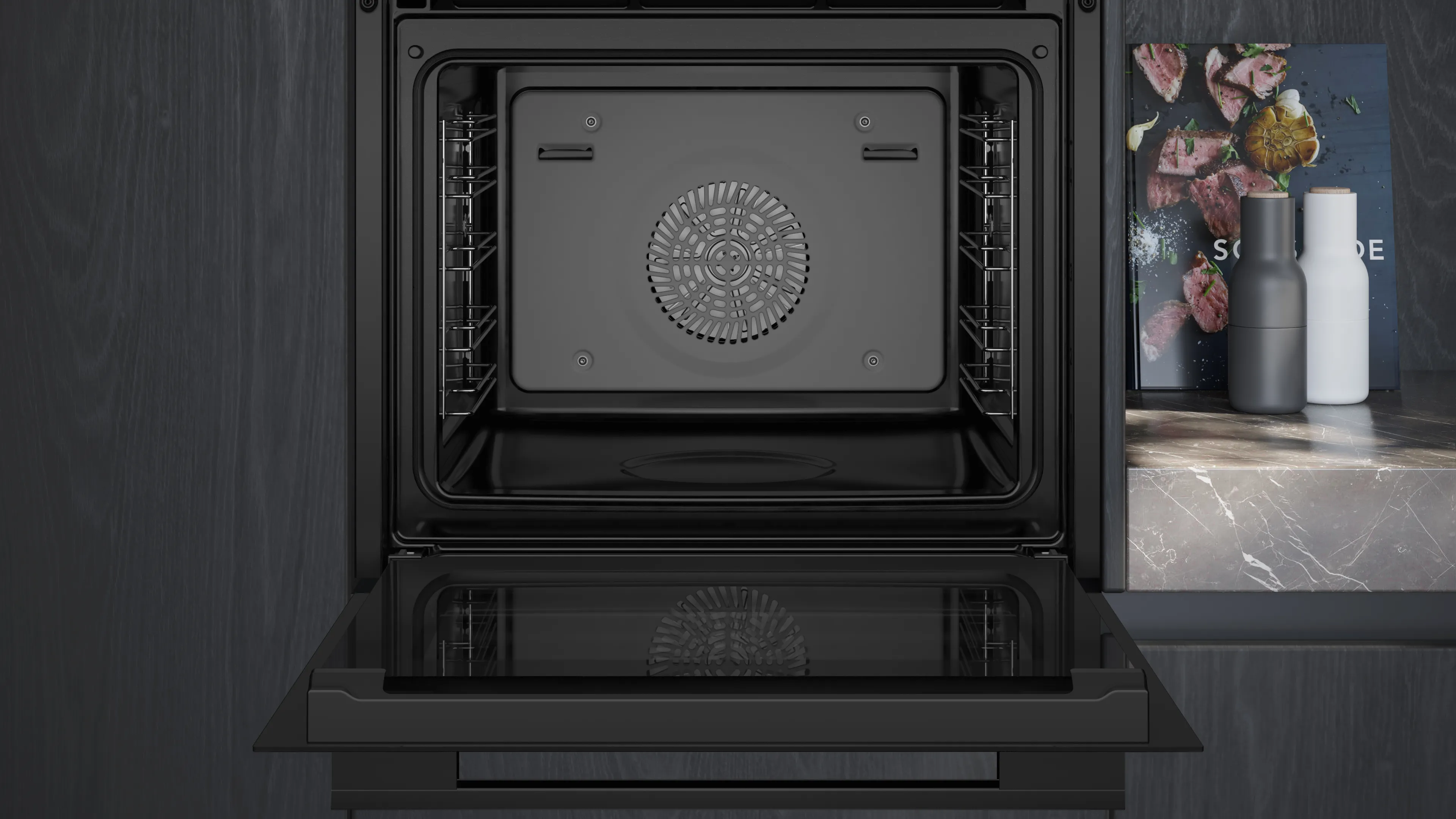 Un horno negro abierto con rejillas internas y ventilador. Un libro de cocina y saleros/pimenteros están sobre la encimera a la derecha.