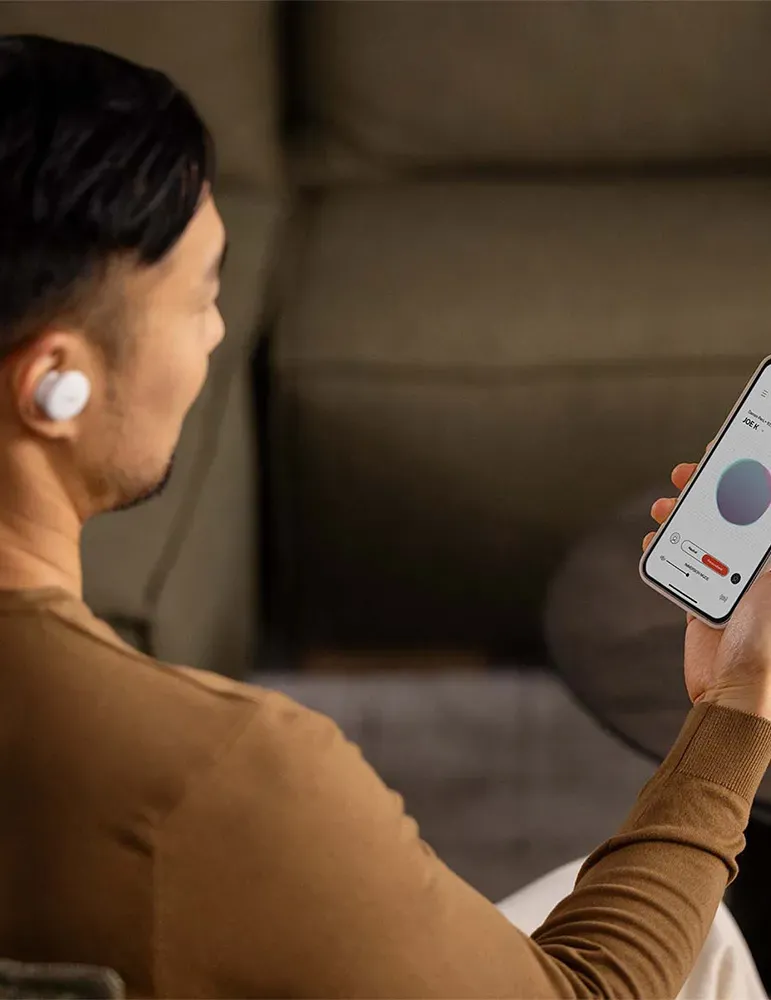 Mężczyzna w słuchawkach, trzymający telefon z interfejsem aplikacji, prawdopodobnie do kontroli dźwięku. Scena jest w pomieszczeniu.