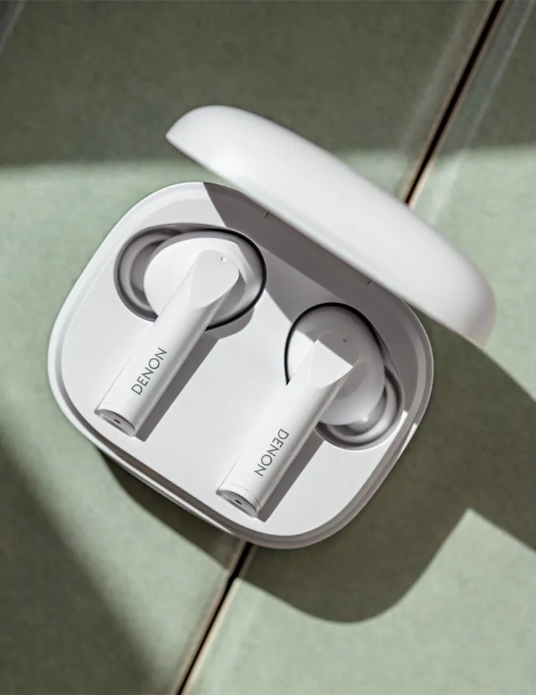 Białe słuchawki Denon w otwartym etui na zielonej powierzchni z płytek. Etui jest białe, a na słuchawkach widnieje napis Denon.