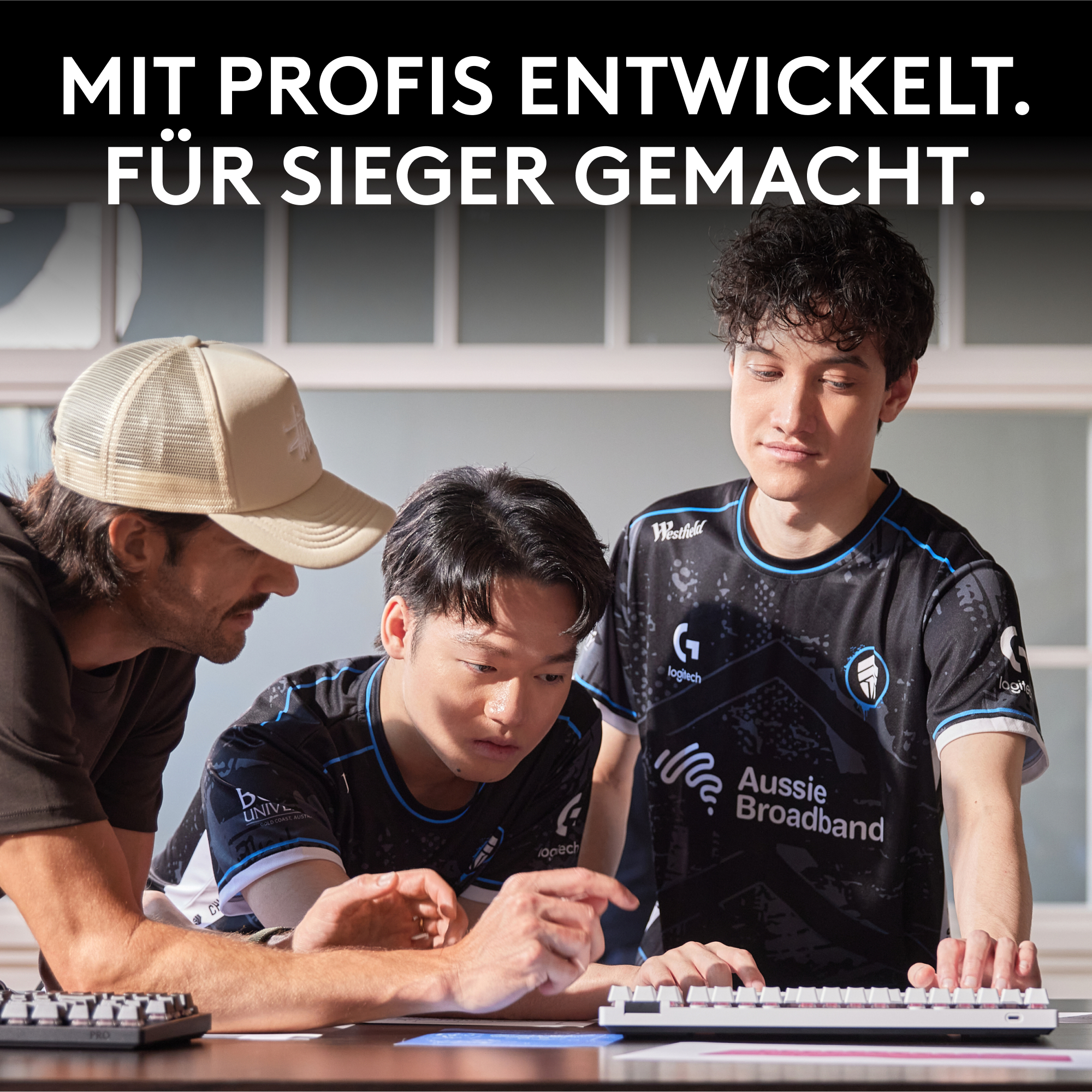 Drei Männer betrachten eine Tastatur. Einer trägt einen Hut, die anderen tragen E-Sport-Trikots. Oben steht der Text 'Für Sieger gemacht'.