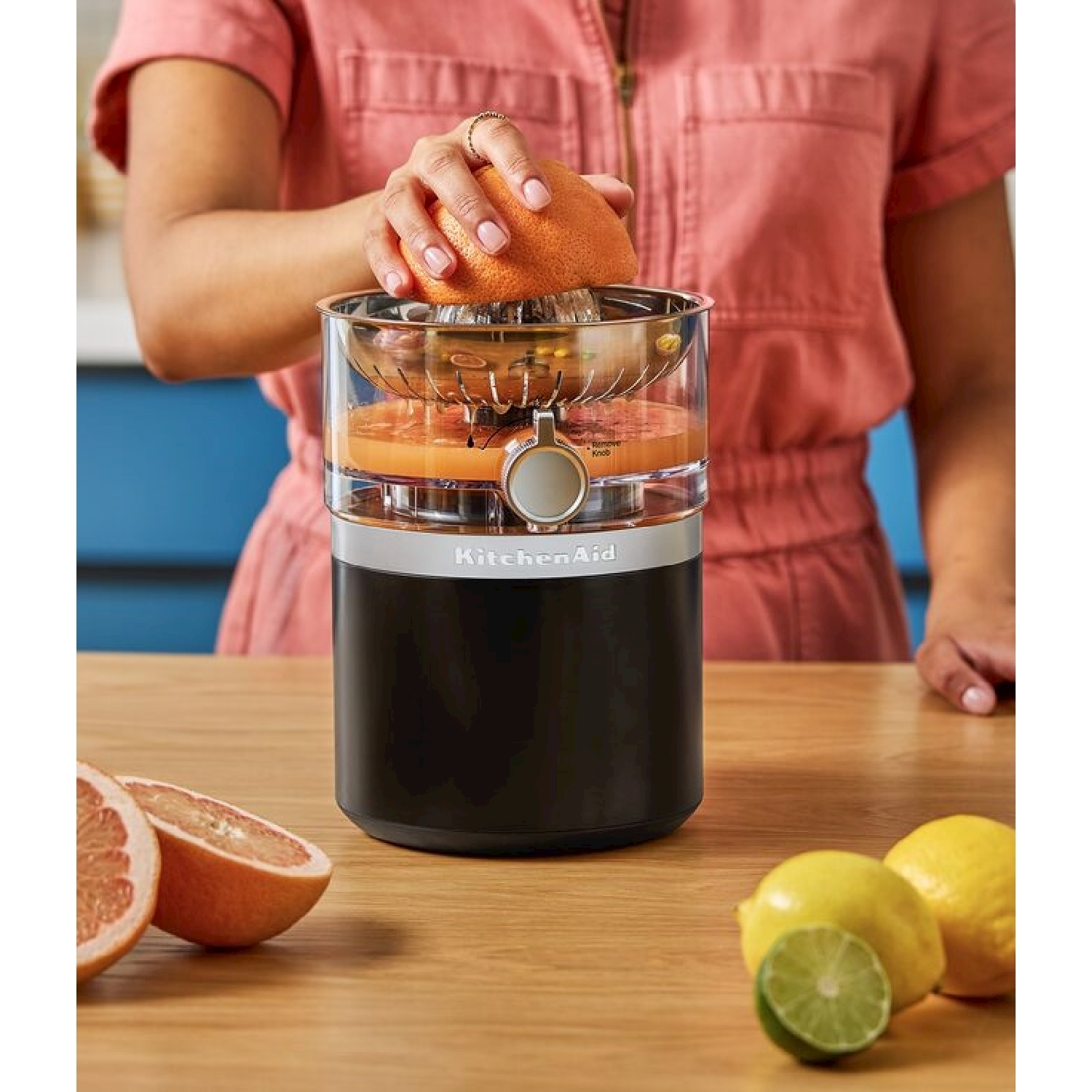 Una persona che sprema un pompelmo con uno spremiagrumi KitchenAid. Pompelmi, limoni e lime tagliati su un tavolo di legno.