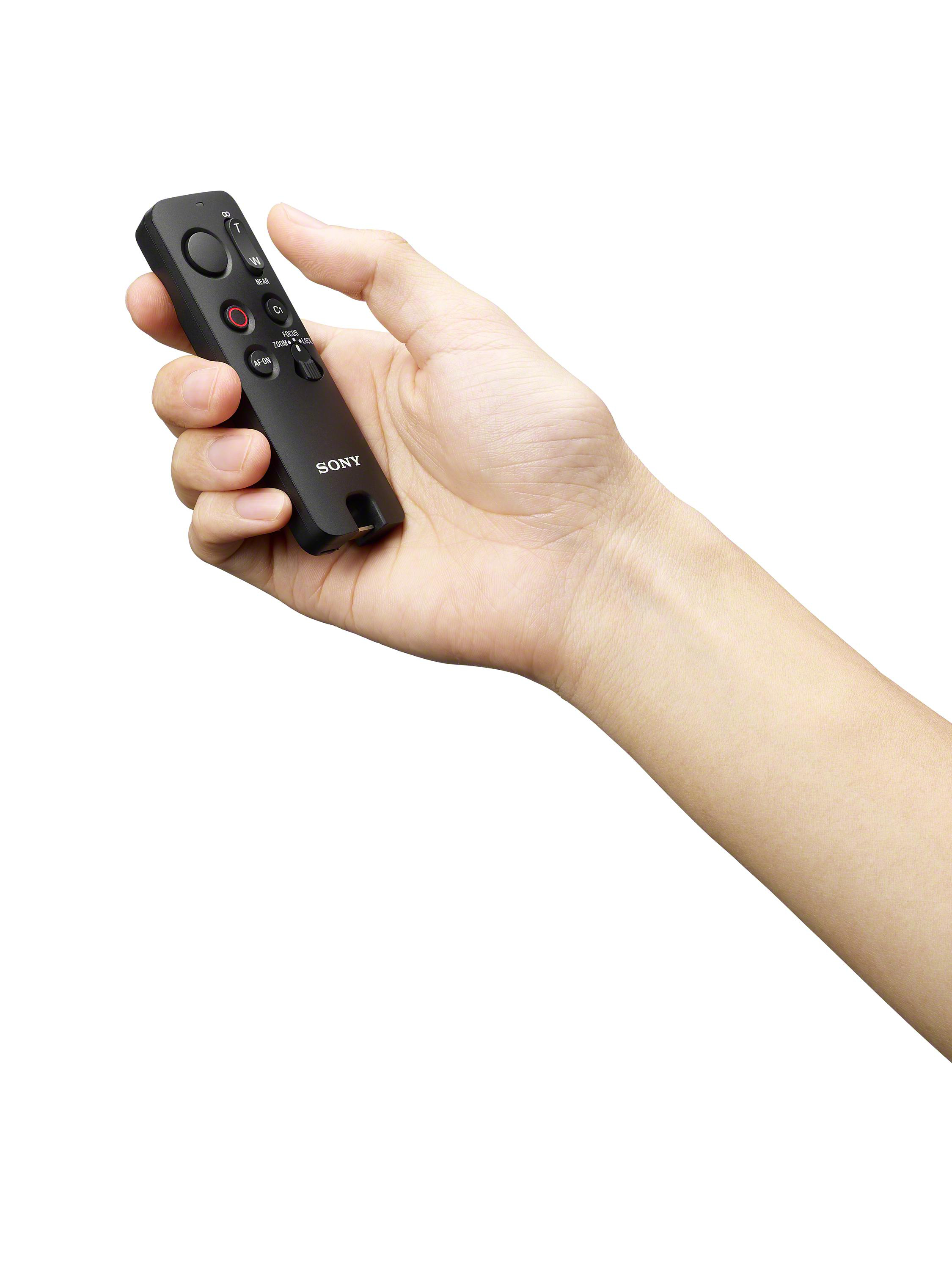 Eine Hand hält eine schwarze Sony-Fernbedienung. Die Fernbedienung hat Knöpfe und einen Schalter für Fokus und Zoom. Der Hintergrund ist weiß.