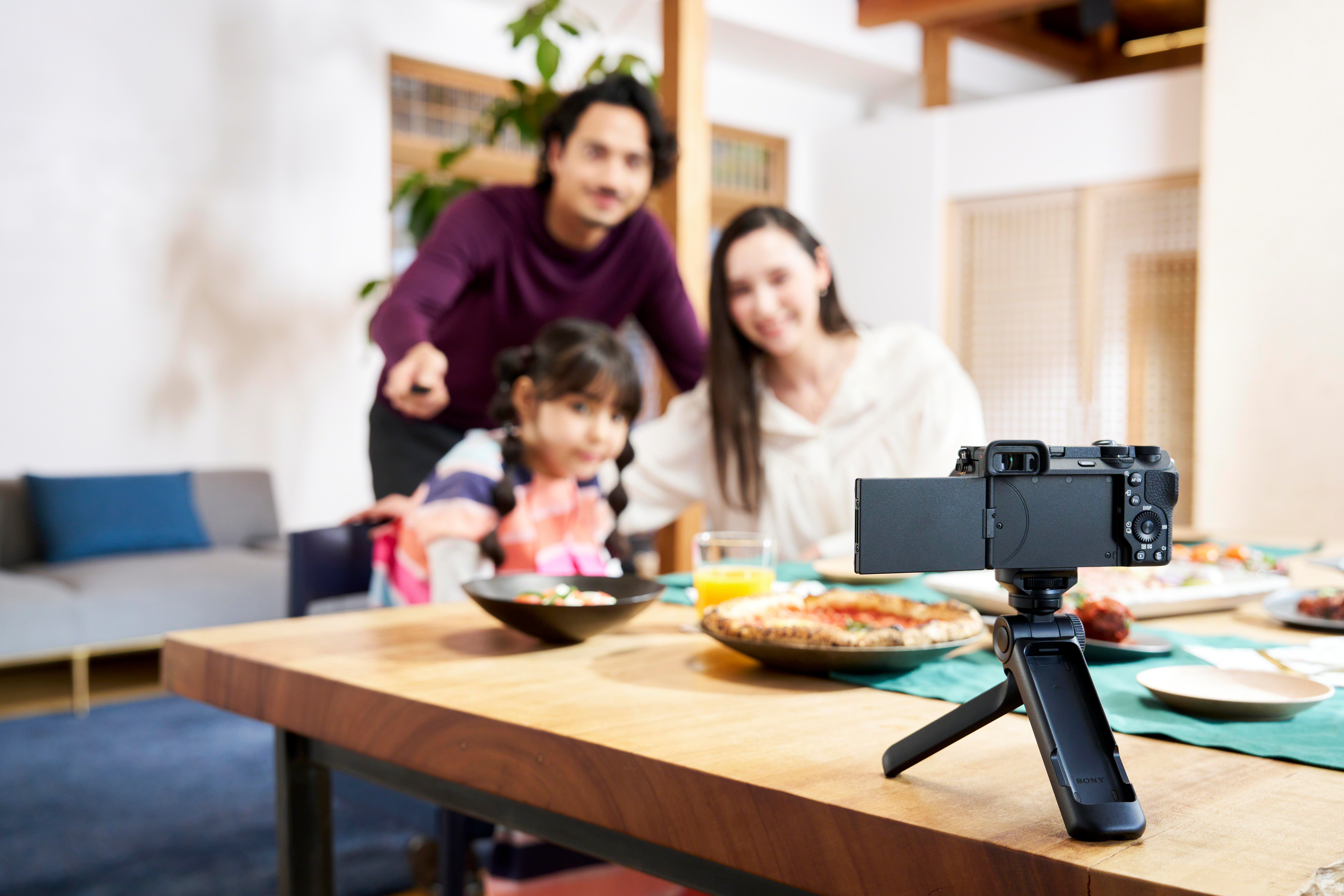 Familie filmt mit einer Kamera auf einem Stativ am Esstisch. Essen und Getränke stehen auf dem Tisch. Im Hintergrund lächelt eine Familie.