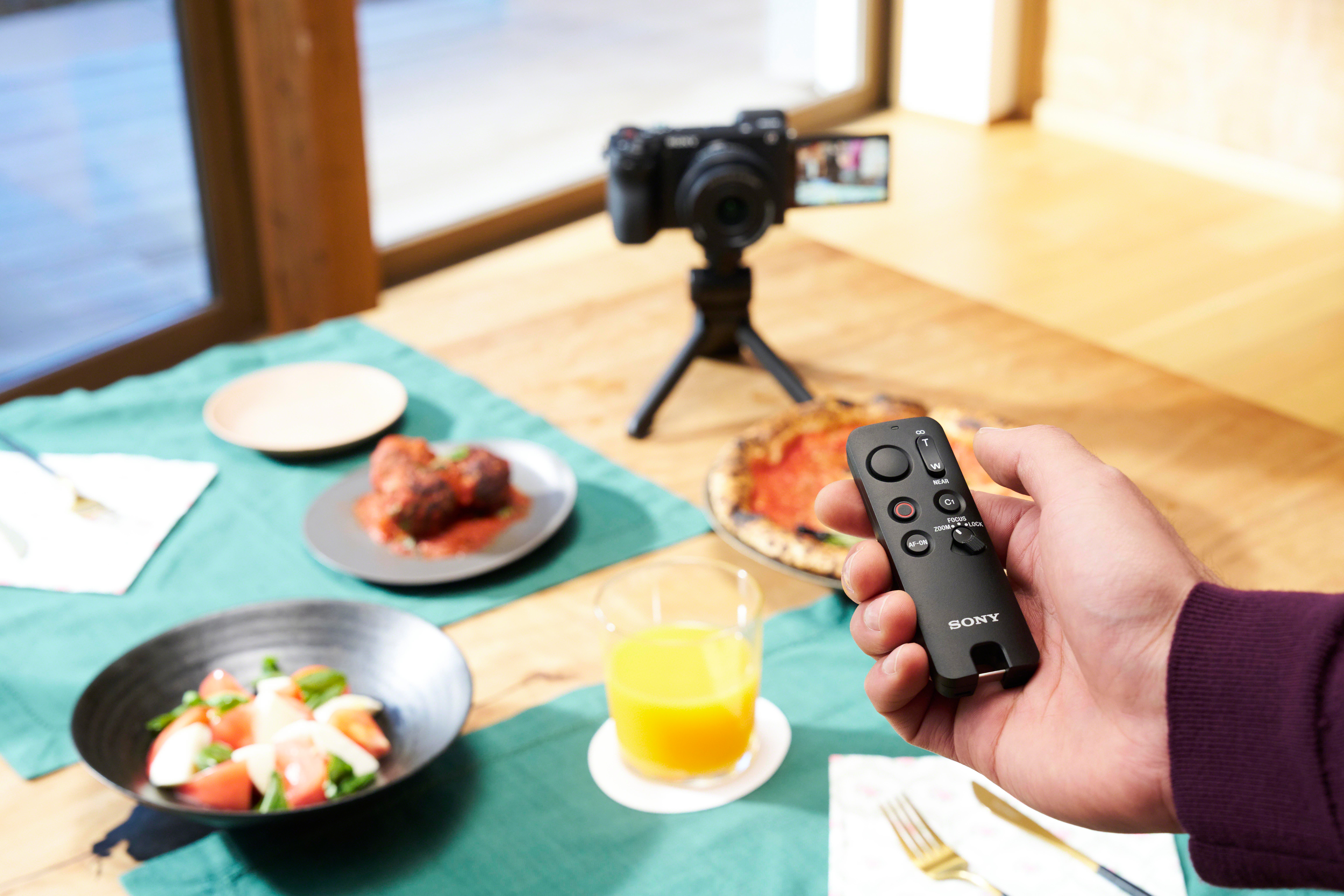 Eine Hand hält eine Sony-Fernbedienung. Eine Kamera auf einem Stativ filmt Essen. Pizza, Salat, Fleischbällchen und Saft liegen auf einem grünen Tischtuch.