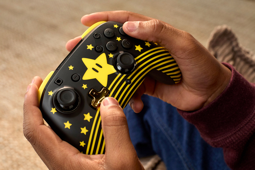 Manos sostienen un mando de juego negro y amarillo con temática de Mario. Una estrella y rayas amarillas lo decoran. El fondo está borroso.