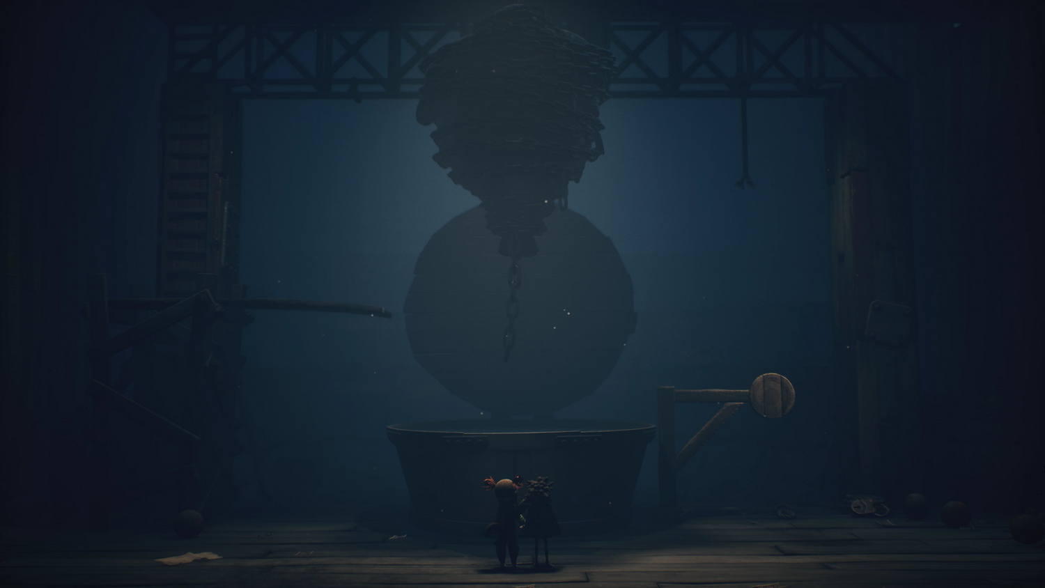 Dos figuras frente a una estructura gigante esférica en un entorno industrial oscuro. La estructura tiene una cadena.