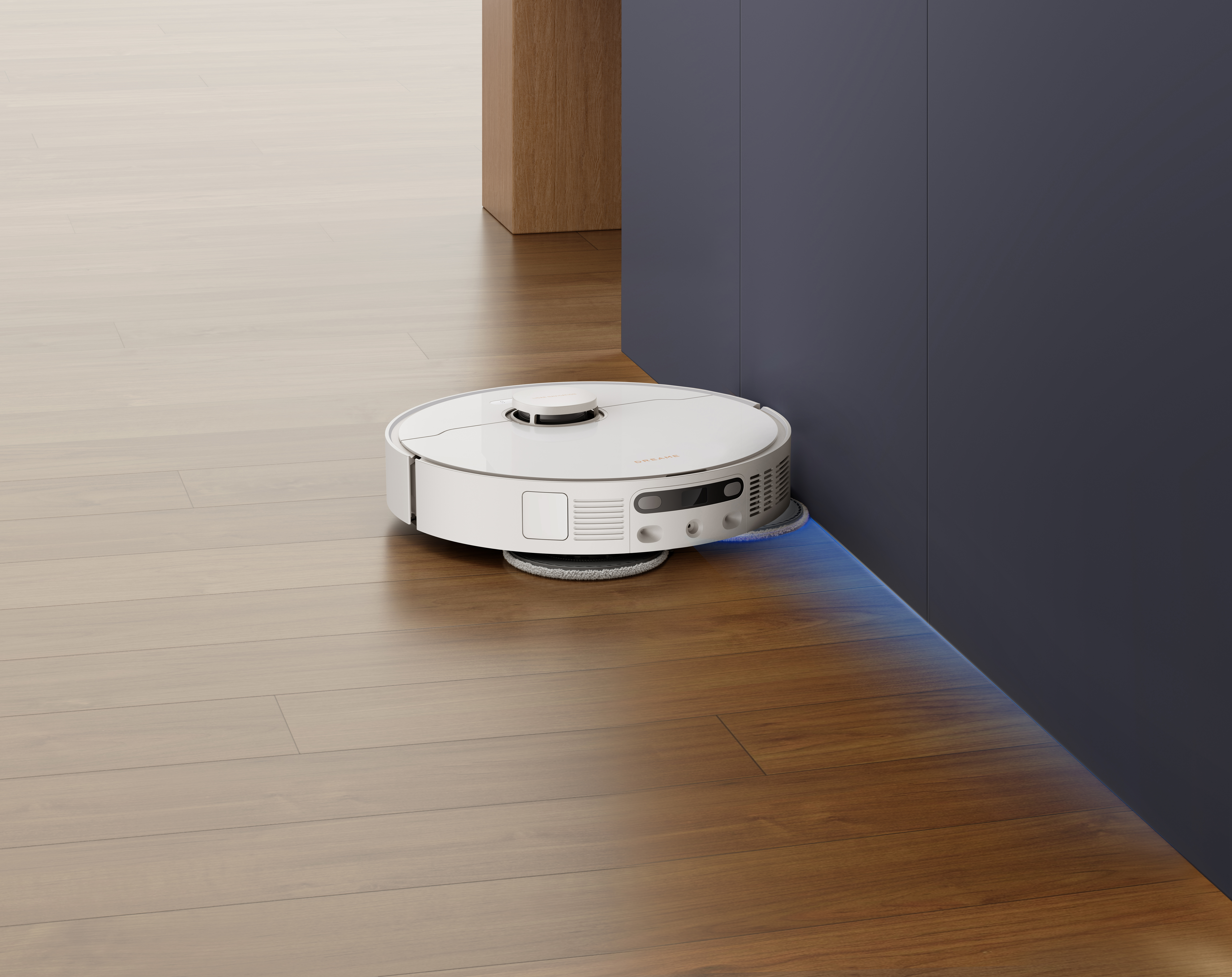 Biały robot odkurzający czyści drewnianą podłogę. Jest blisko ciemnoniebieskiej ściany i drewnianej szafki.