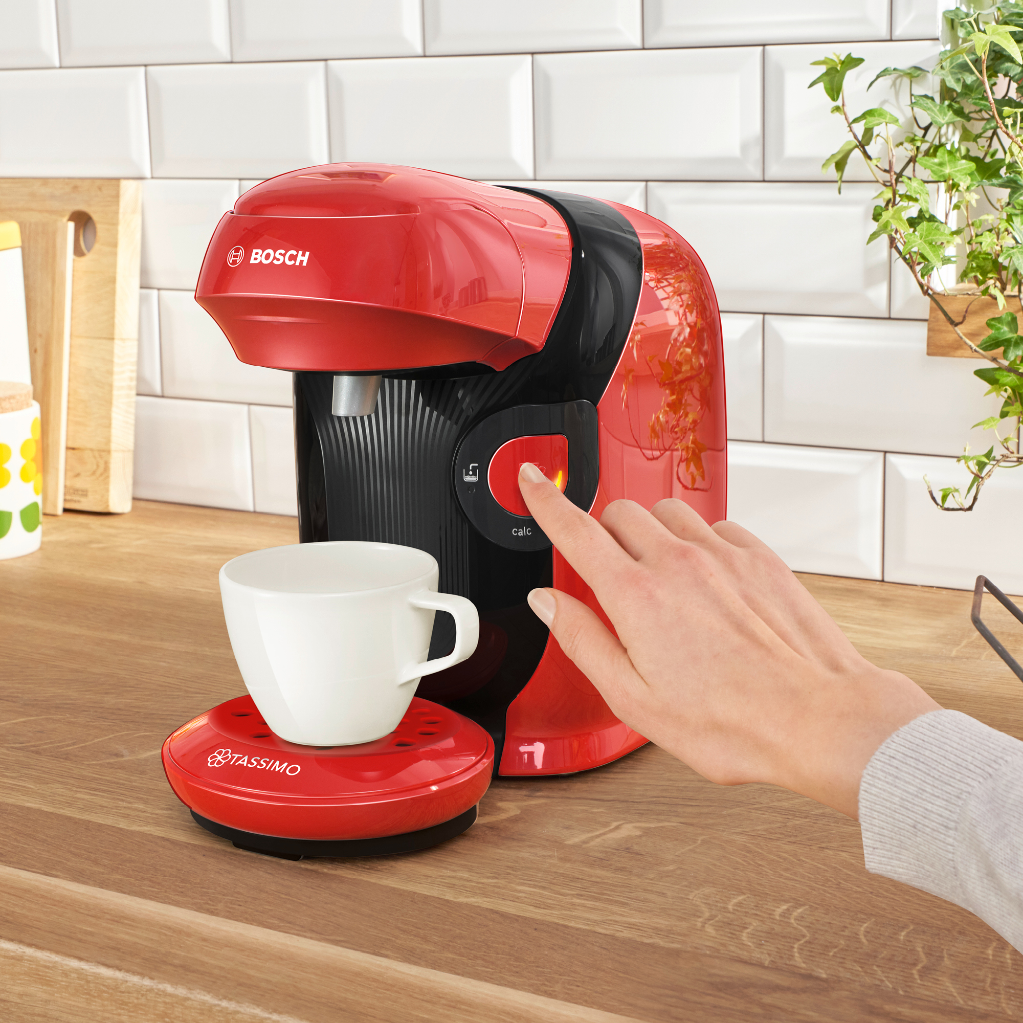 Cafetera roja y negra con una taza blanca sobre una base roja. Una mano pulsa el botón. Azulejos blancos al fondo.