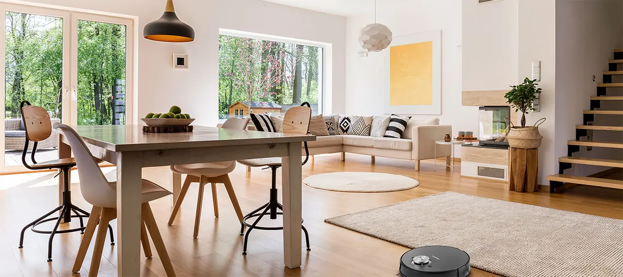 Modern nappali. Asztal székekkel, kanapé, szőnyeg és robotporszívó. Ablak a fákra nyíló kilátással.