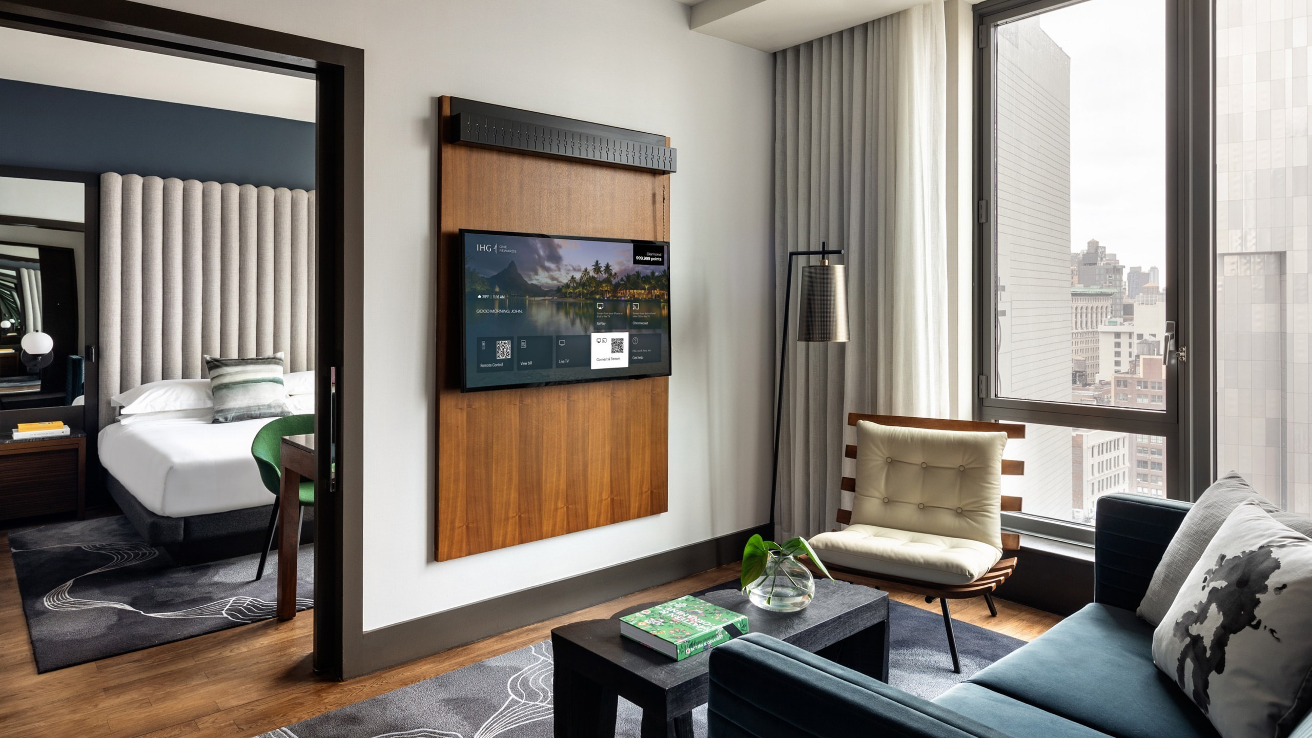 Şehir manzaralı modern bir otel odası. Ahşap bir panelde TV, bir kanepe, bir sandalye ve bir kapıdan görülebilen bir yatak bulunmaktadır.