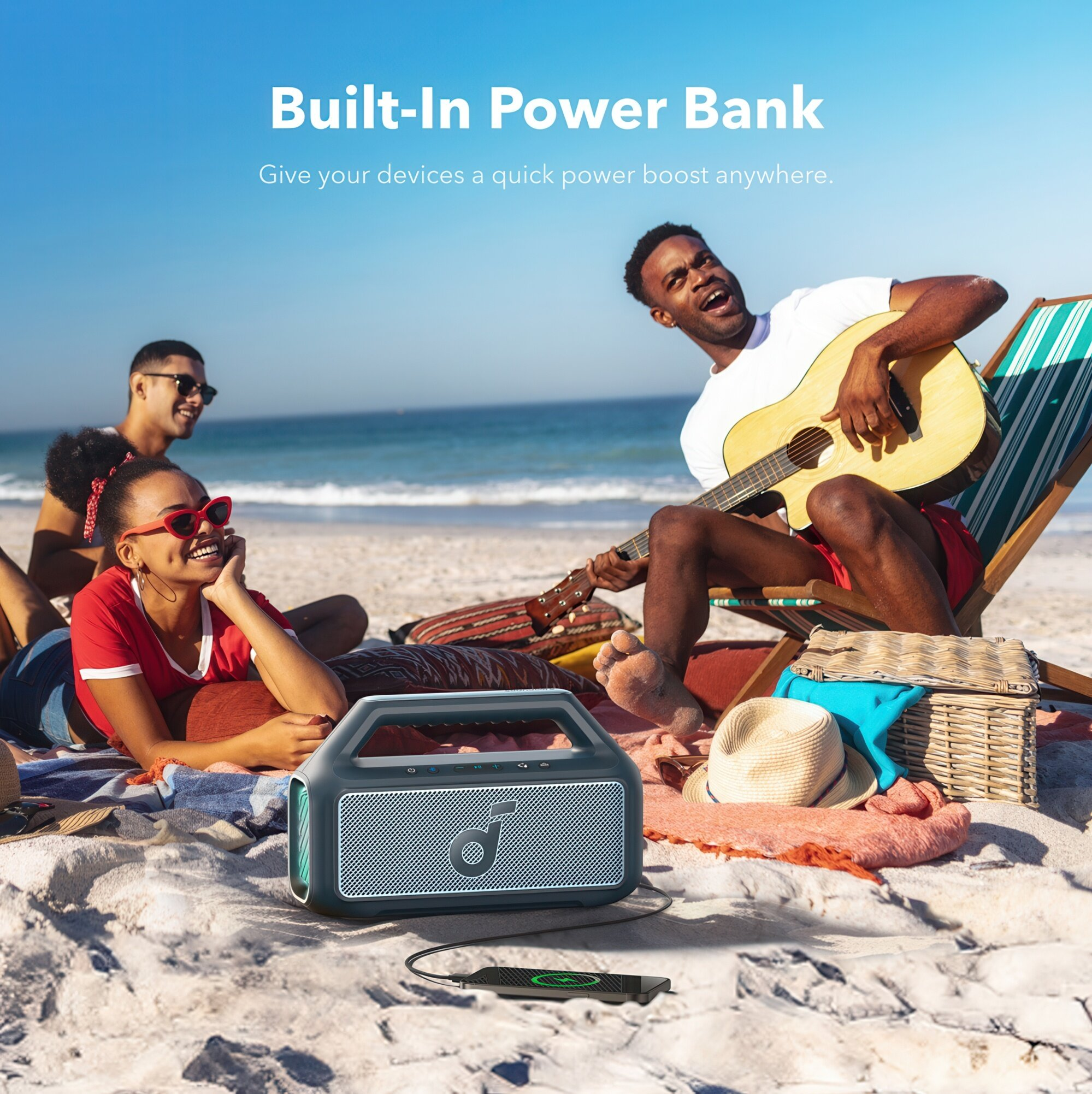 Ludzie na plaży. Jeden gra na gitarze, inni odpoczywają. Głośnik ładuje telefon. Błękitne niebo i morze w tle.