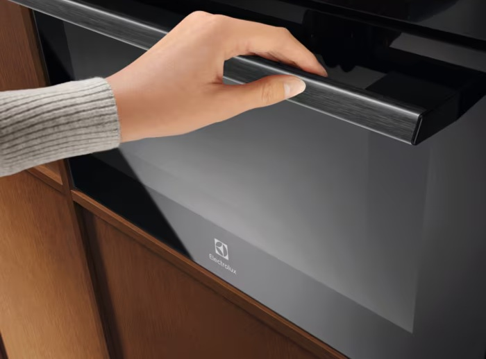 Ręka otwiera czarne drzwi piekarnika z uchwytem ze szczotkowanego metalu. Piekarnik jest wbudowany w drewnianą szafkę.