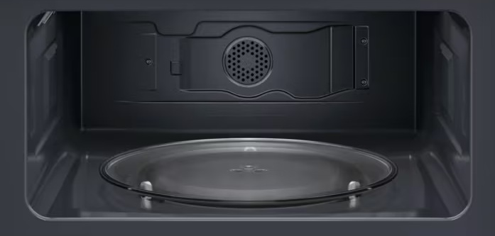 Wnętrze kuchenki mikrofalowej: szklany talerz na dole, ciemne wnętrze z otworami wentylacyjnymi i elementami na górze.