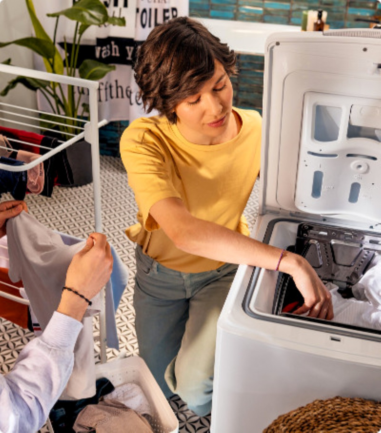 Kobieta wkłada pranie do pralki. Inna osoba podaje jej ubrania. Kosz na pranie jest na pierwszym planie. Suszarka znajduje się po lewej stronie.