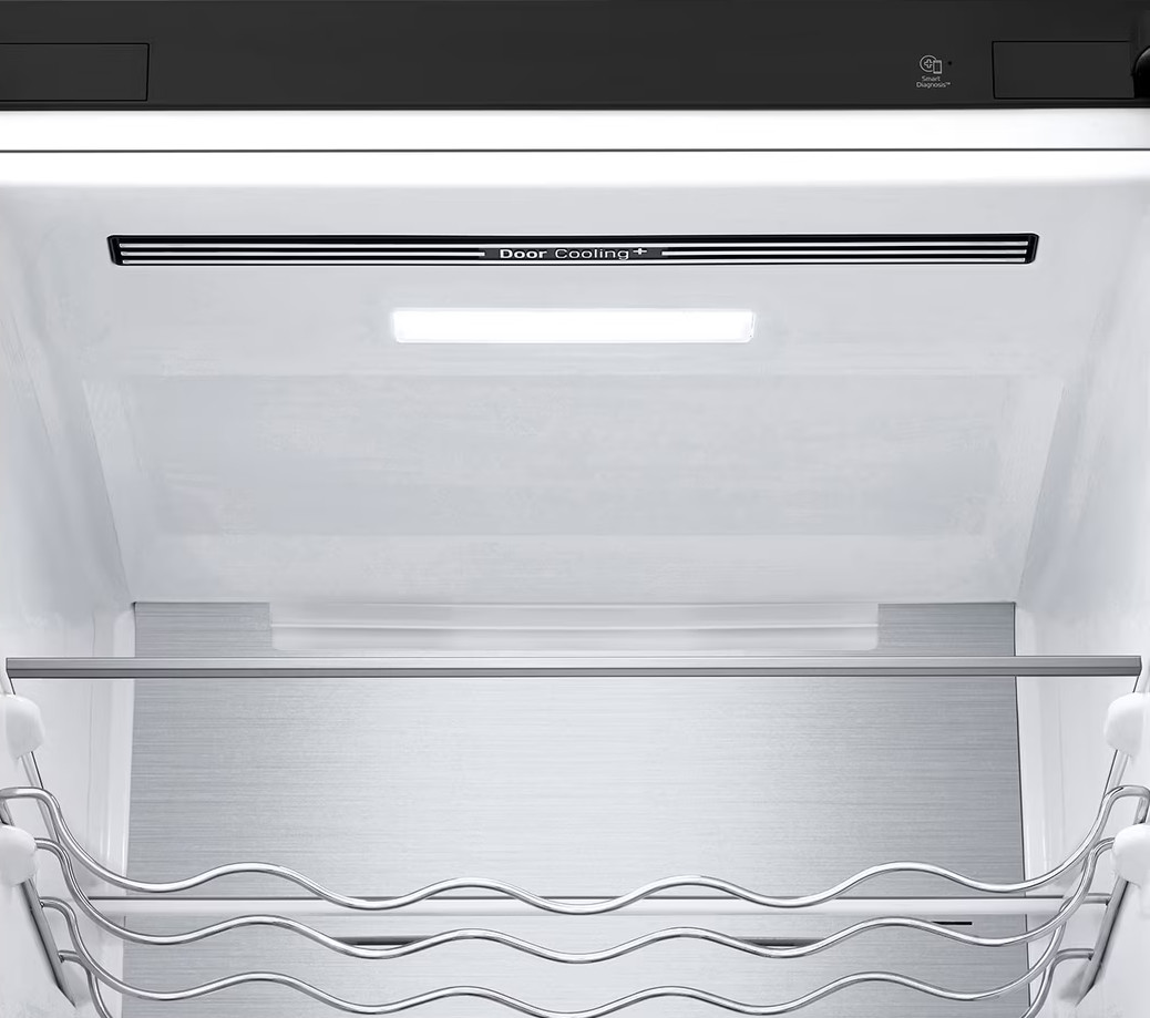 Wewnątrz lodówki. Światło świeci do środka. Są półki i stojaki na jedzenie i napoje. Ściany są białe.