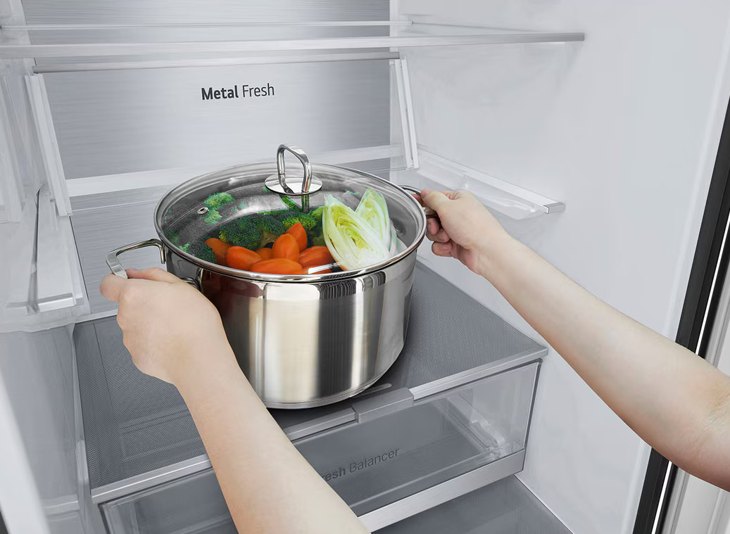 Ręce trzymające garnek z warzywami w lodówce. Widoczny brokuł, marchew i sałata.