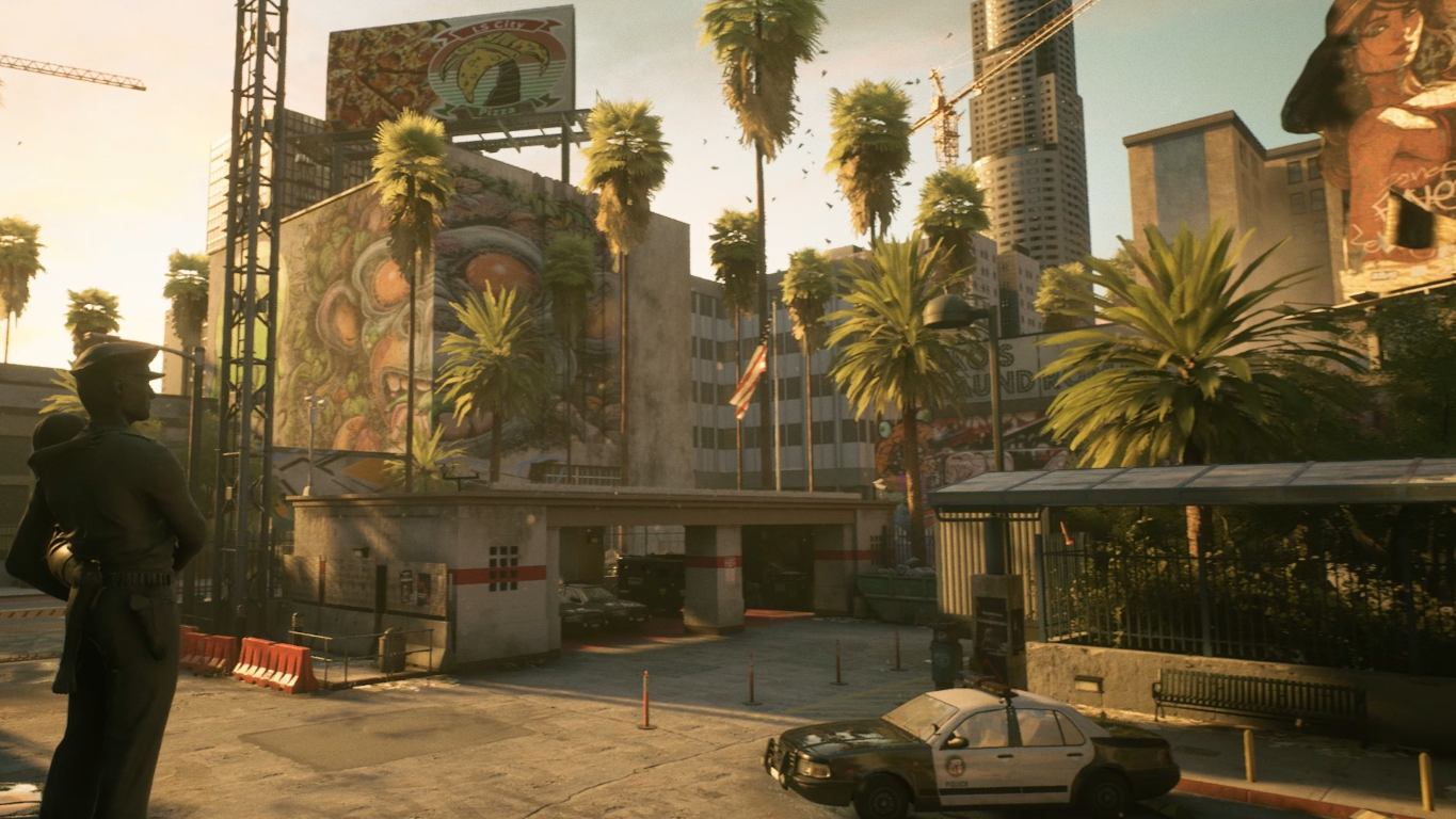 Pejzaż miejski z palmami, budynkami, muralami i radiowozem. Na pierwszym planie stoi posąg osoby trzymającej dziecko.