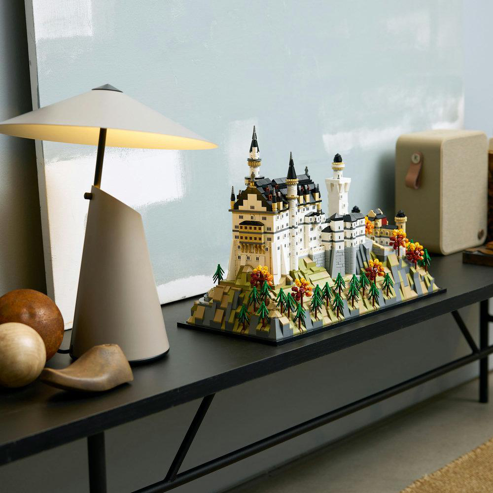 Ein Lego-Modell von Schloss Neuschwanstein steht auf einem schwarzen Regal mit Lampe und Dekoration. Das Schloss ist weiß mit schwarzen Dächern.