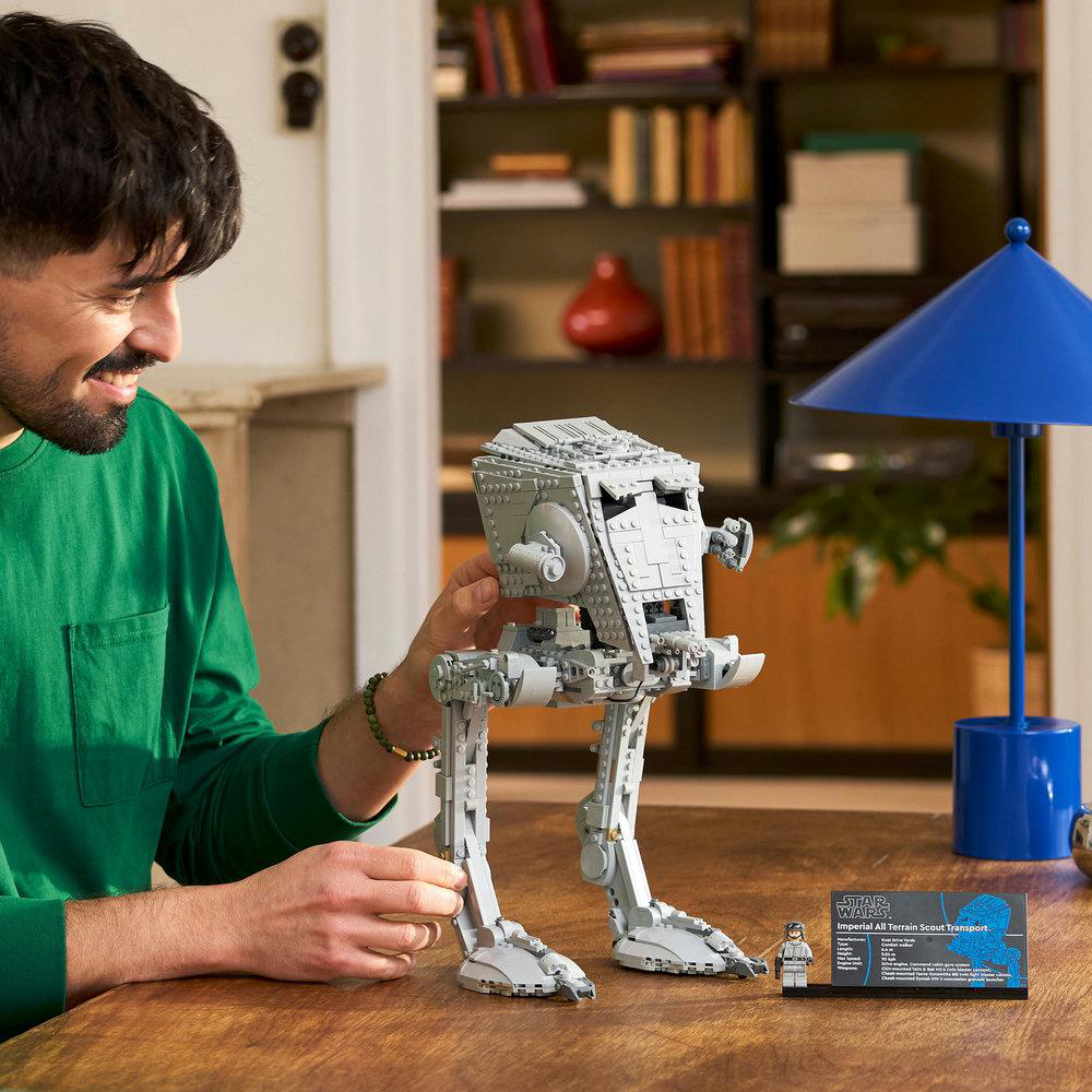 Ein Mann lächelt und hält einen Lego AT-ST. Rechts ist eine blaue Lampe, auf dem Tisch eine Star Wars Karte. Im Hintergrund ein Bücherregal.
