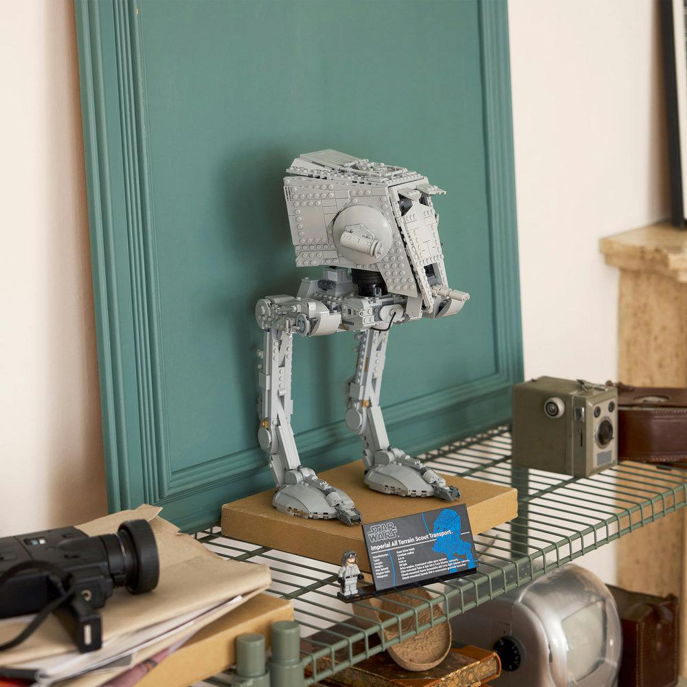 Ein Lego AT-ST-Modell steht auf einem Regal. Dahinter ist eine grüne Wand. Auf dem Regal befinden sich weitere Gegenstände, darunter eine Kamera und Bücher.