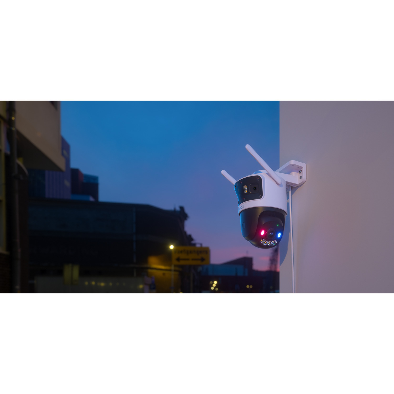 Een witte beveiligingscamera met antennes is aan een muur bevestigd. De achtergrond toont gebouwen en een straat in de schemering.