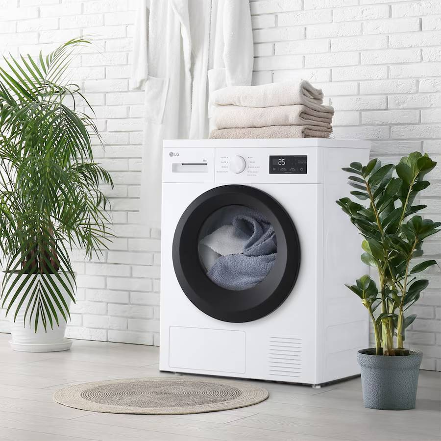 Una secadora blanca con puerta negra en una habitación. Toallas apiladas encima. Plantas y una alfombra en primer plano.