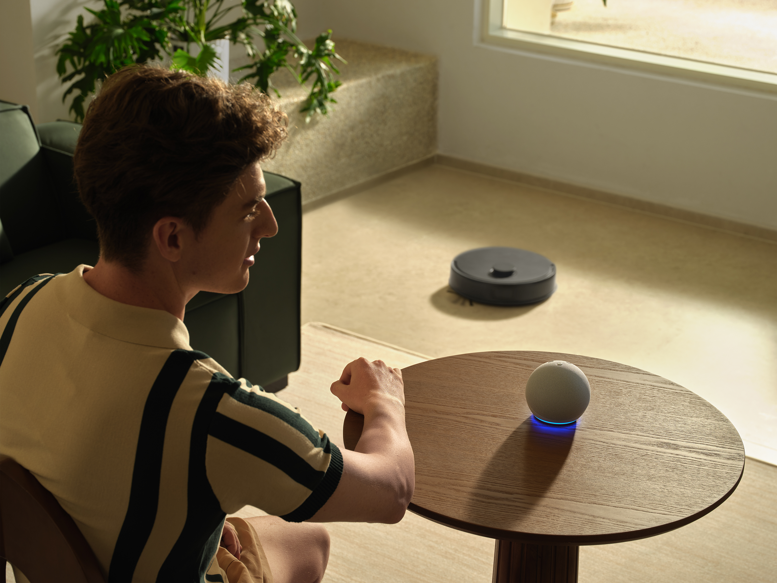 Egy férfi egy okos hangszóróval ellátott asztal közelében ül. Egy robotporszívó a háttérben látható.