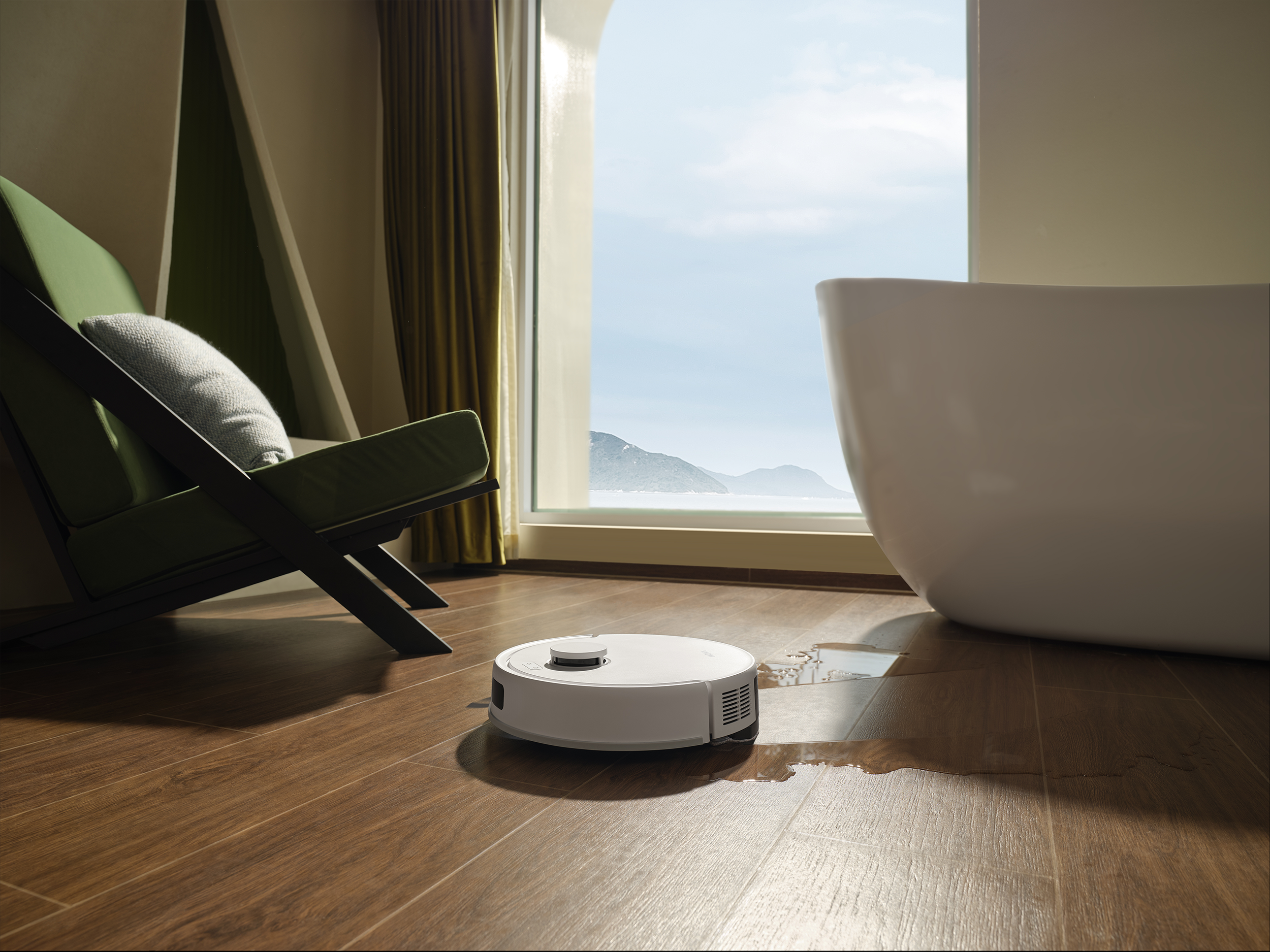 Fehér robotporszívó a fa padlón, egy fehér fürdőkád és egy zöld szék közelében, hegyek a háttérben.