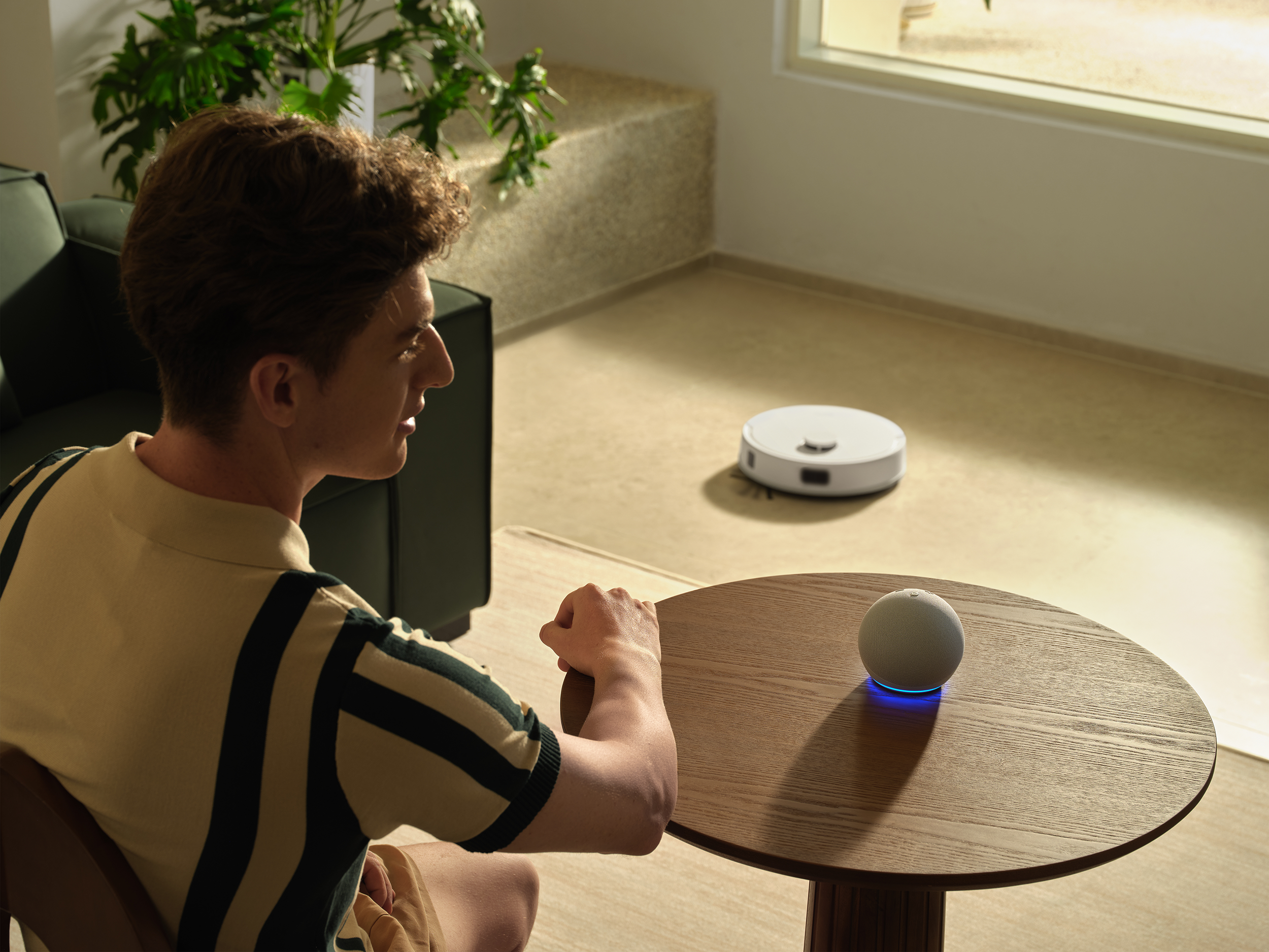 Egy férfi egy asztalnál ül okoshangszóróval. Egy robotporszívó a padlón van. Növények és egy ablak a háttérben.