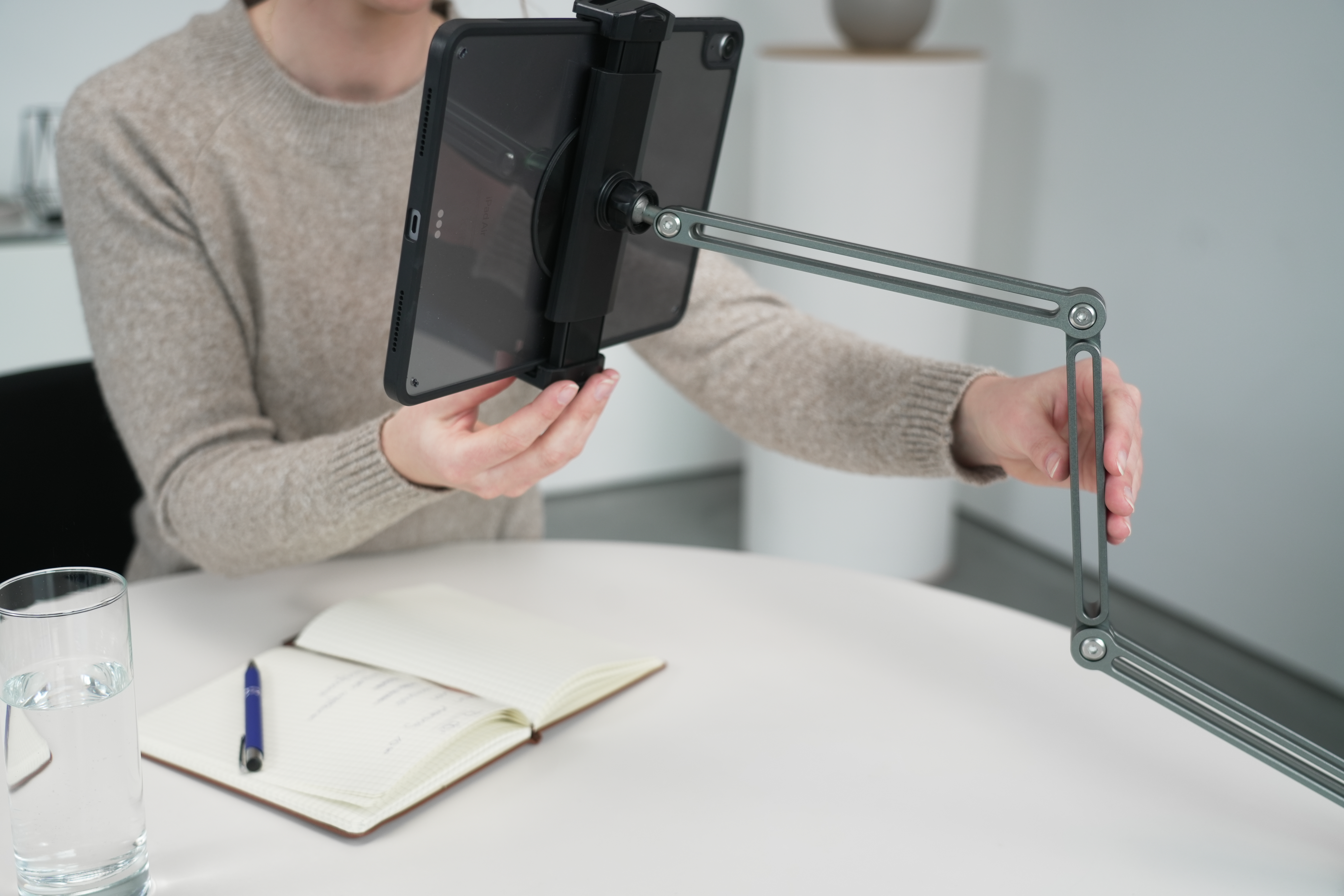 Eine Person hält ein Tablet an einem grauen Arm, ein Notizbuch, Stift und ein Glas Wasser liegen auf dem Tisch.