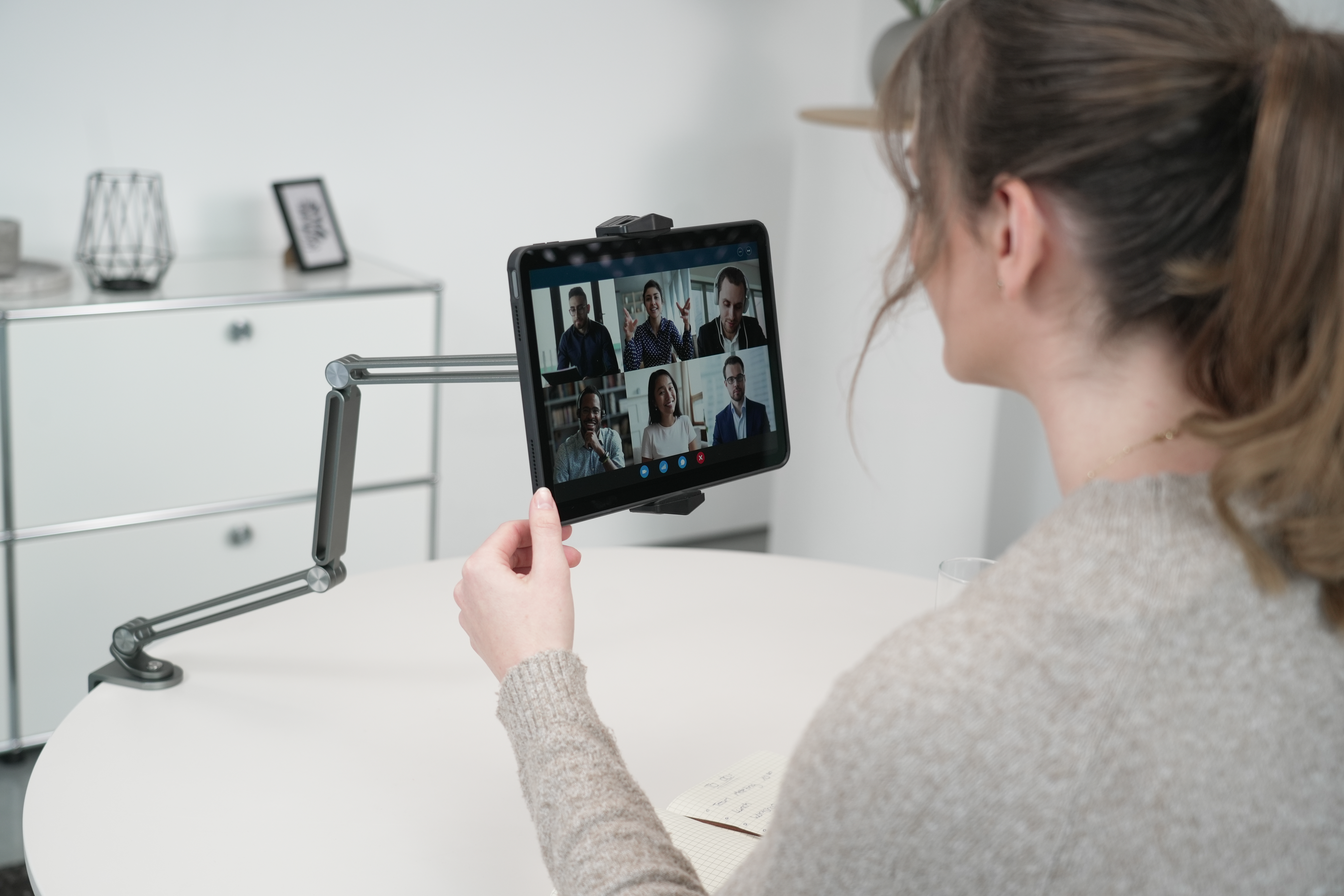 Eine Frau nimmt an einem Videoanruf auf einem Tablet teil. Das Tablet ist montiert und zeigt mehrere Teilnehmer an. Ein Notizblock ist ebenfalls vorhanden.