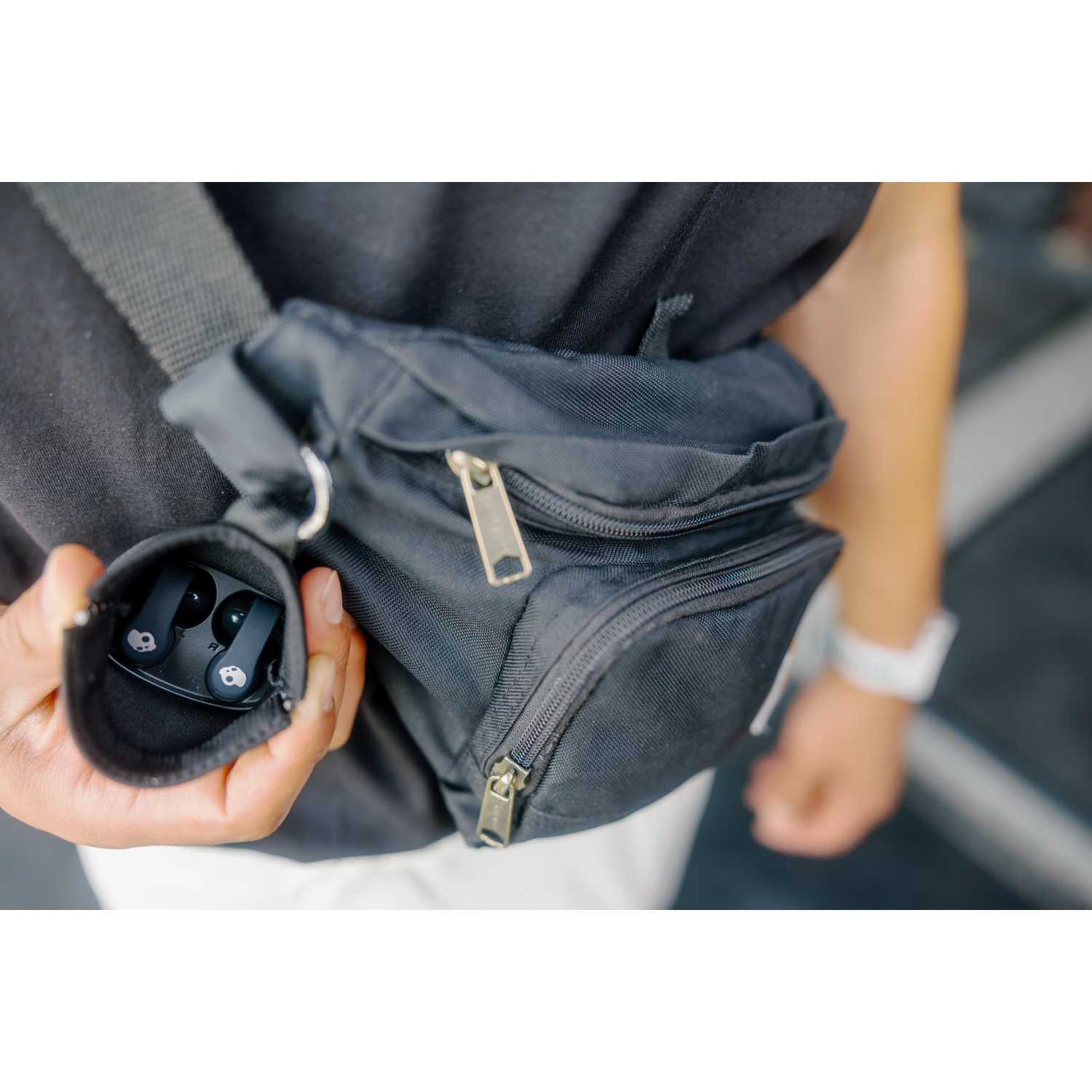 Personne tenant des écouteurs noirs dans un étui, avec un sac noir. Une montre est au poignet. L'arrière-plan est flou.