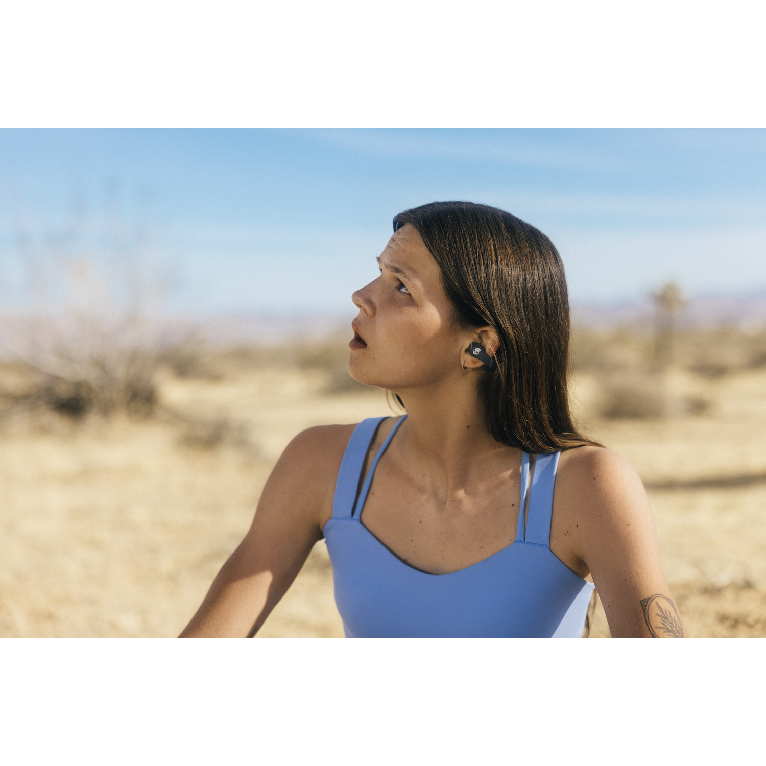 Une femme en haut bleu regarde en l'air, portant des écouteurs. Elle est dans un désert avec un ciel bleu. Un tatouage est visible sur son bras.