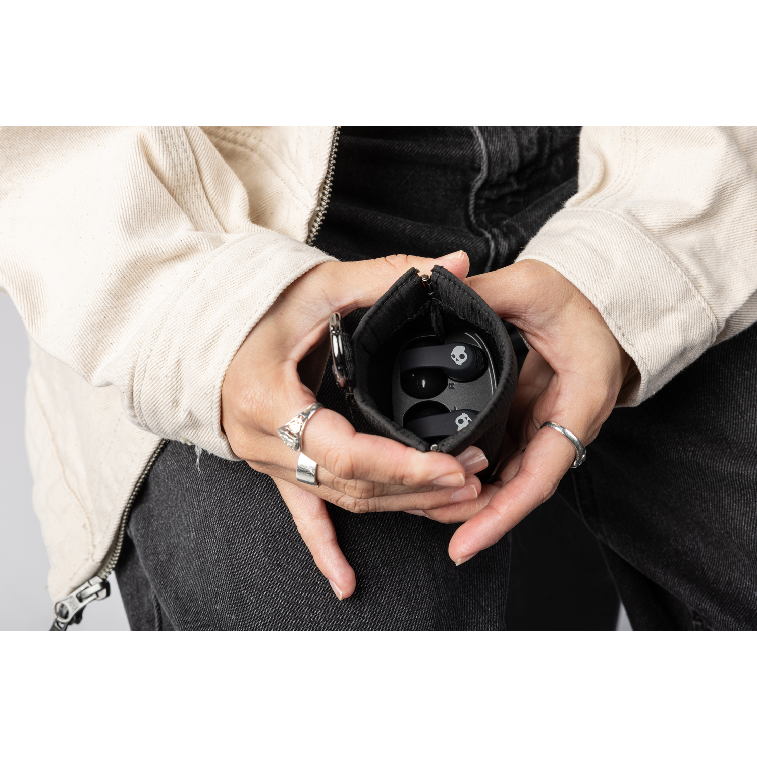 Personne tenant un étui noir avec des écouteurs. Il porte une veste crème et un jean noir. Des bagues ornent ses doigts.