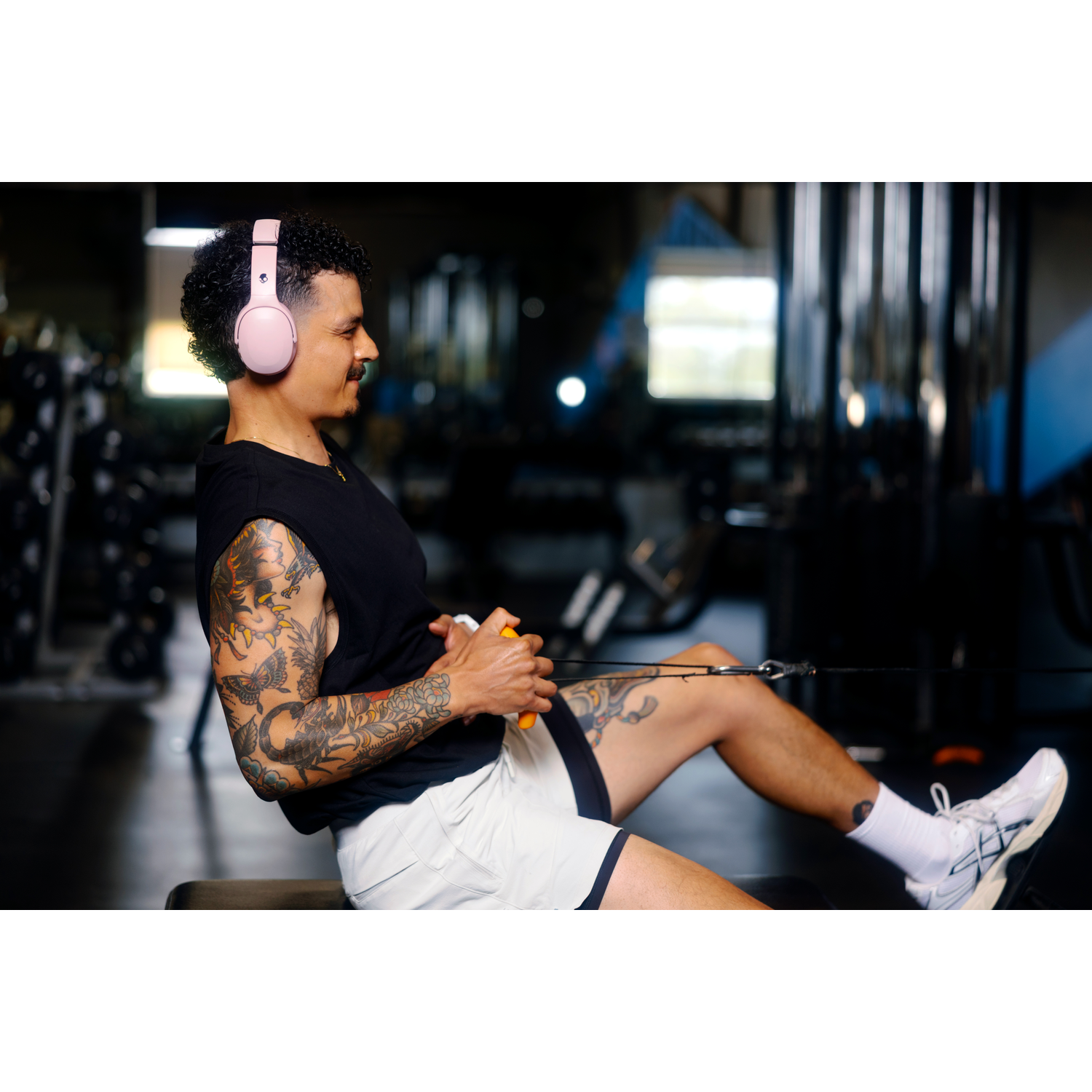 Homme tatoué dans une salle de sport, portant un casque audio. Il utilise un rameur. Il porte un débardeur noir et un short blanc.