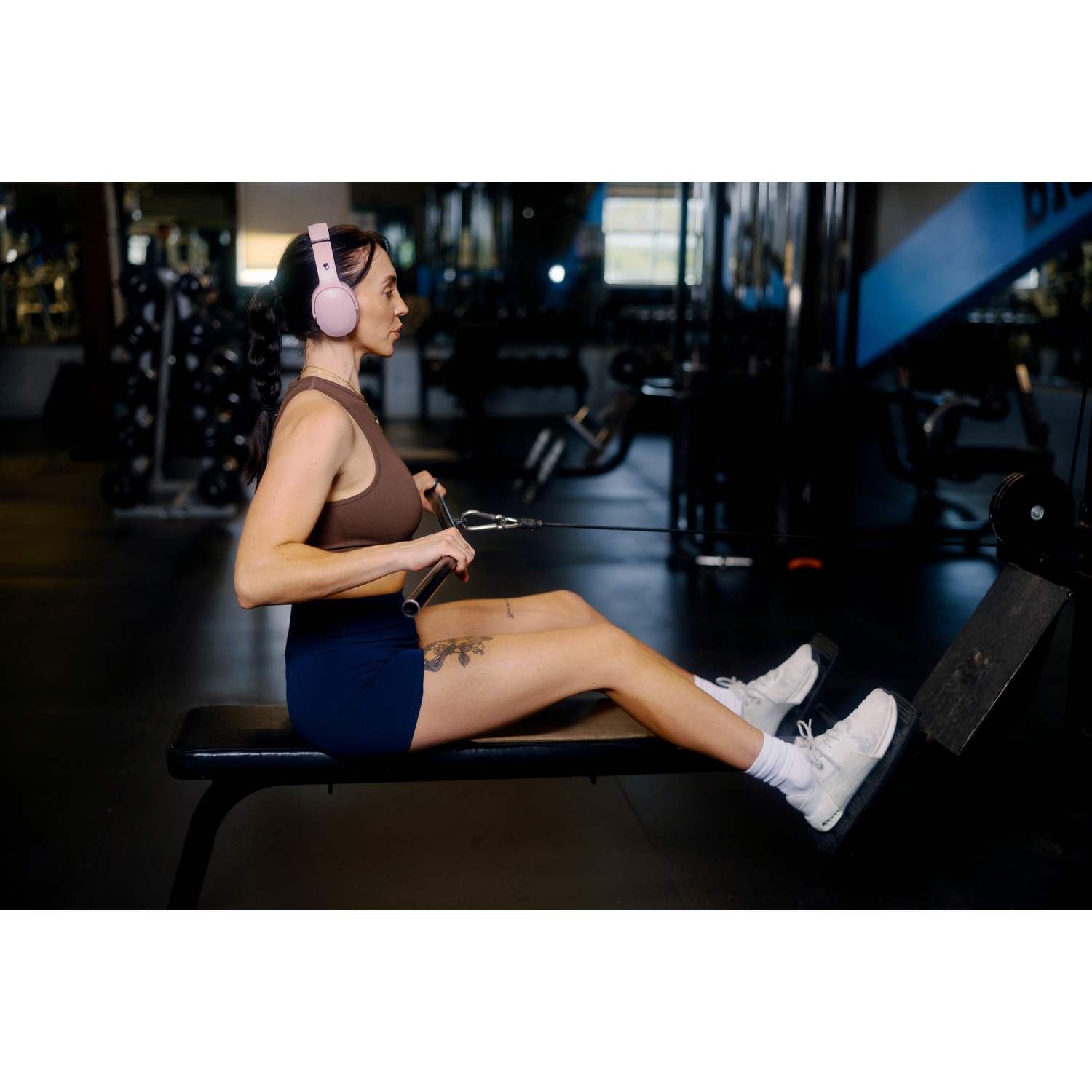 Femme en casque audio faisant de l'aviron sur une machine de gym. Elle porte un haut marron, un short bleu et des chaussures blanches. Équipement de gym en arrière-plan.
