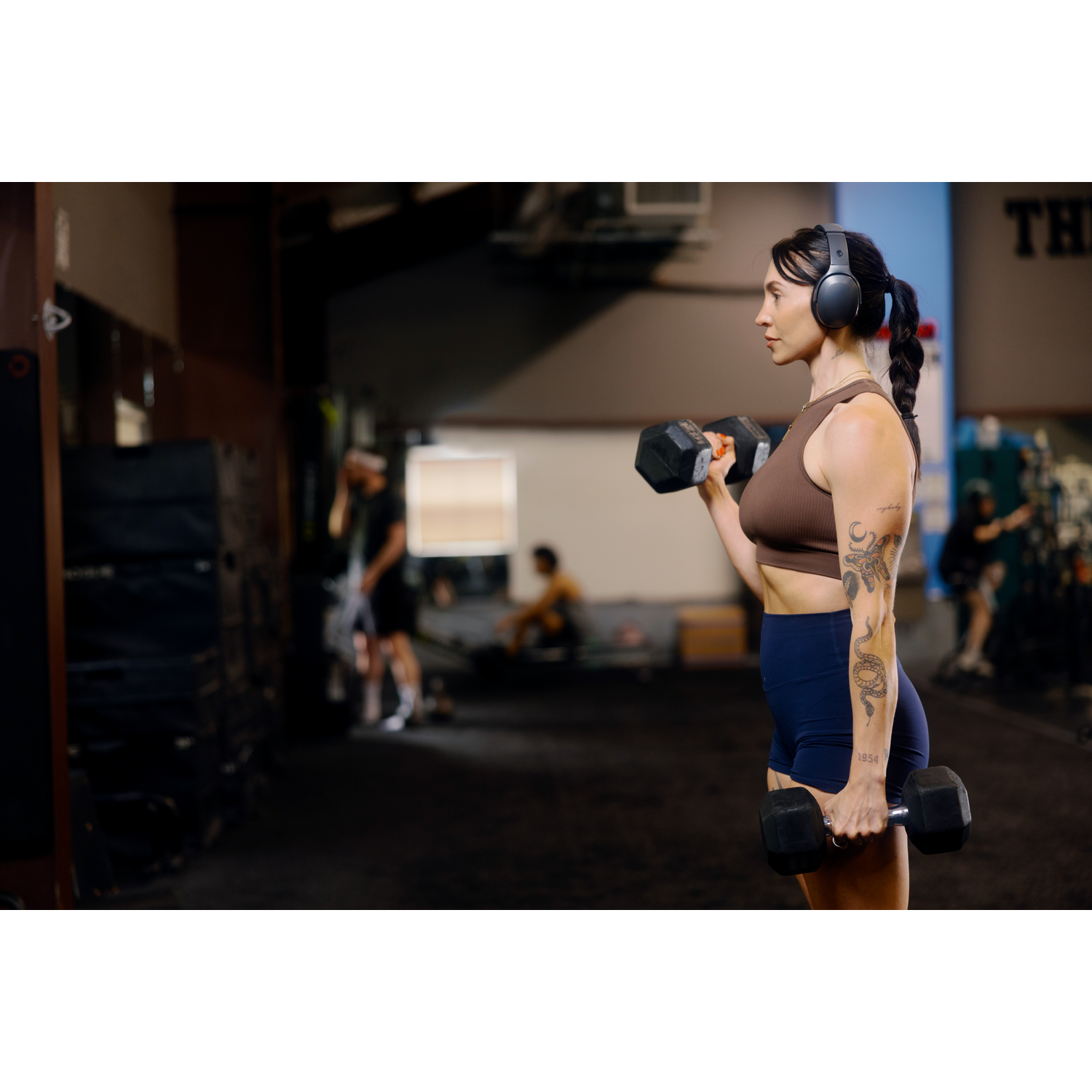 Une femme dans une salle de sport lève des haltères. Elle porte un casque, un haut marron et un short bleu. D'autres personnes et équipements sont en arrière-plan.
