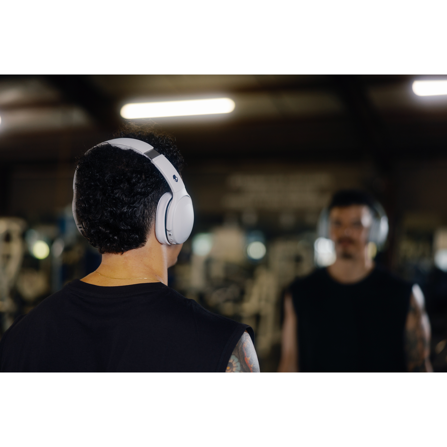 Homme portant un casque blanc dans une salle de sport. Un autre homme reflété en arrière-plan. Les deux portent des hauts noirs.