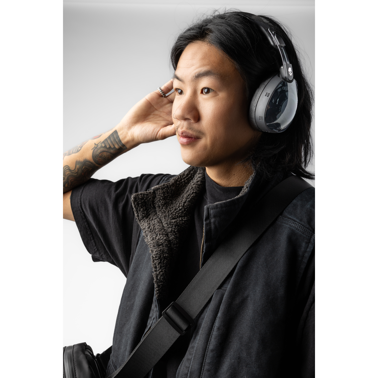 Homme aux cheveux noirs, avec un casque et une veste noire. Il a un tatouage sur le bras. Un sac est sur son épaule.