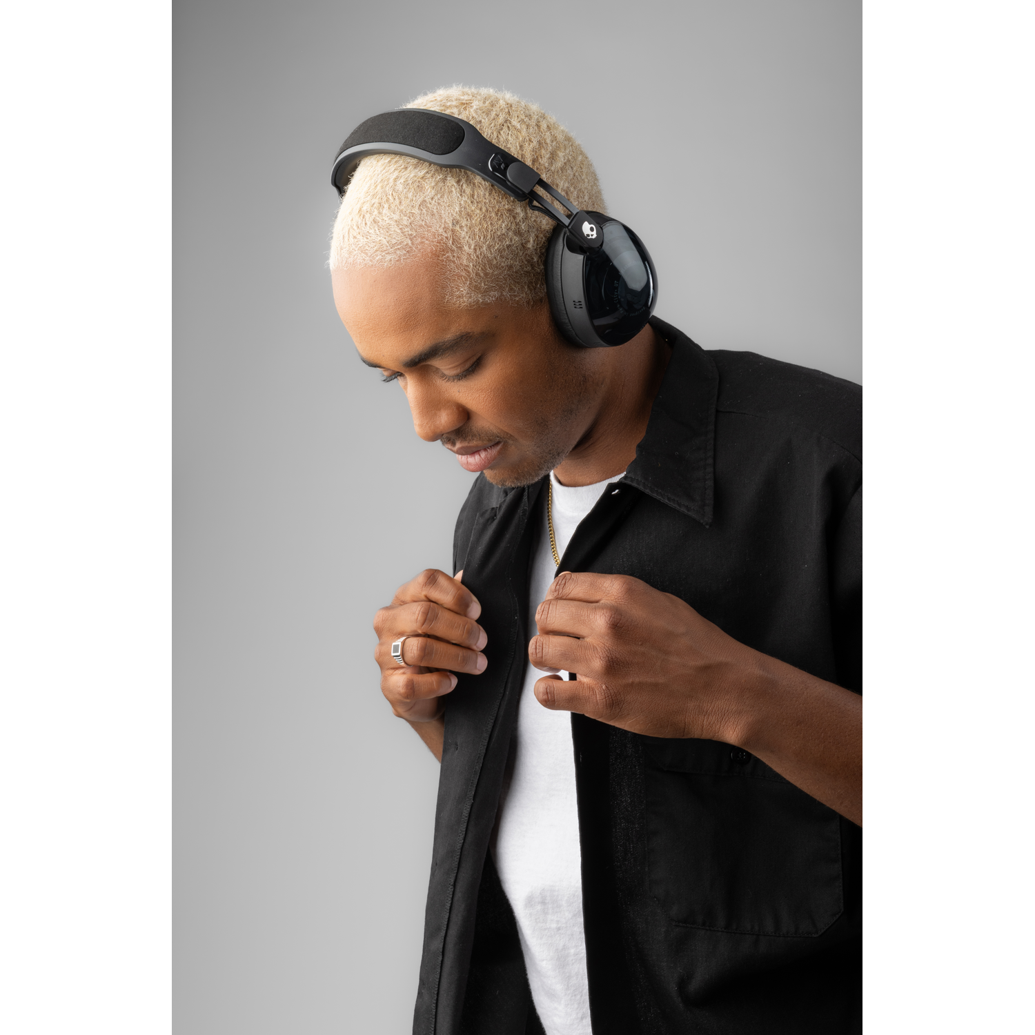 Un homme aux cheveux blonds portant un casque et une chemise noire, regardant vers le bas. Une chaîne en or et une bague sont visibles.