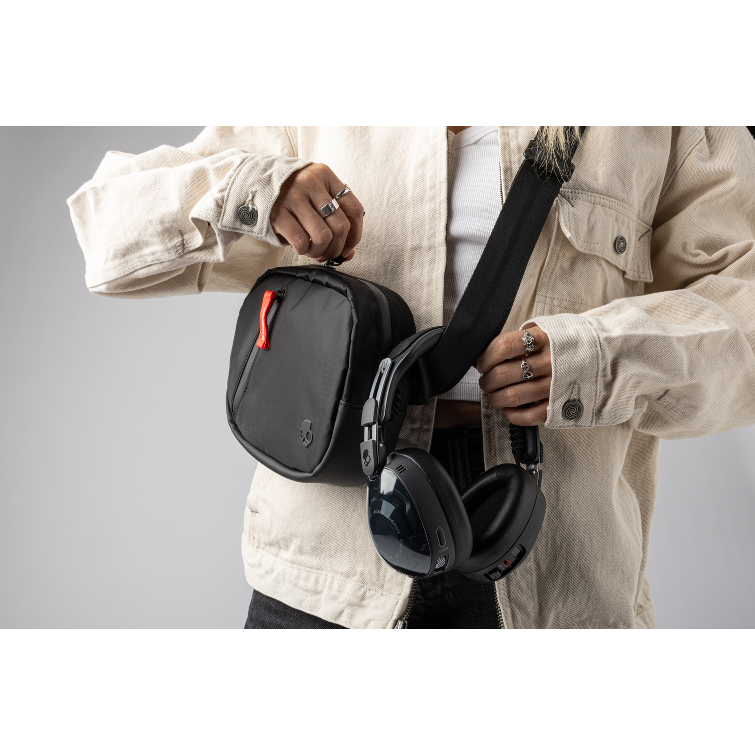 Personne tenant un sac noir et un casque. Elle porte une veste beige et des bagues. Le fond est gris uni.