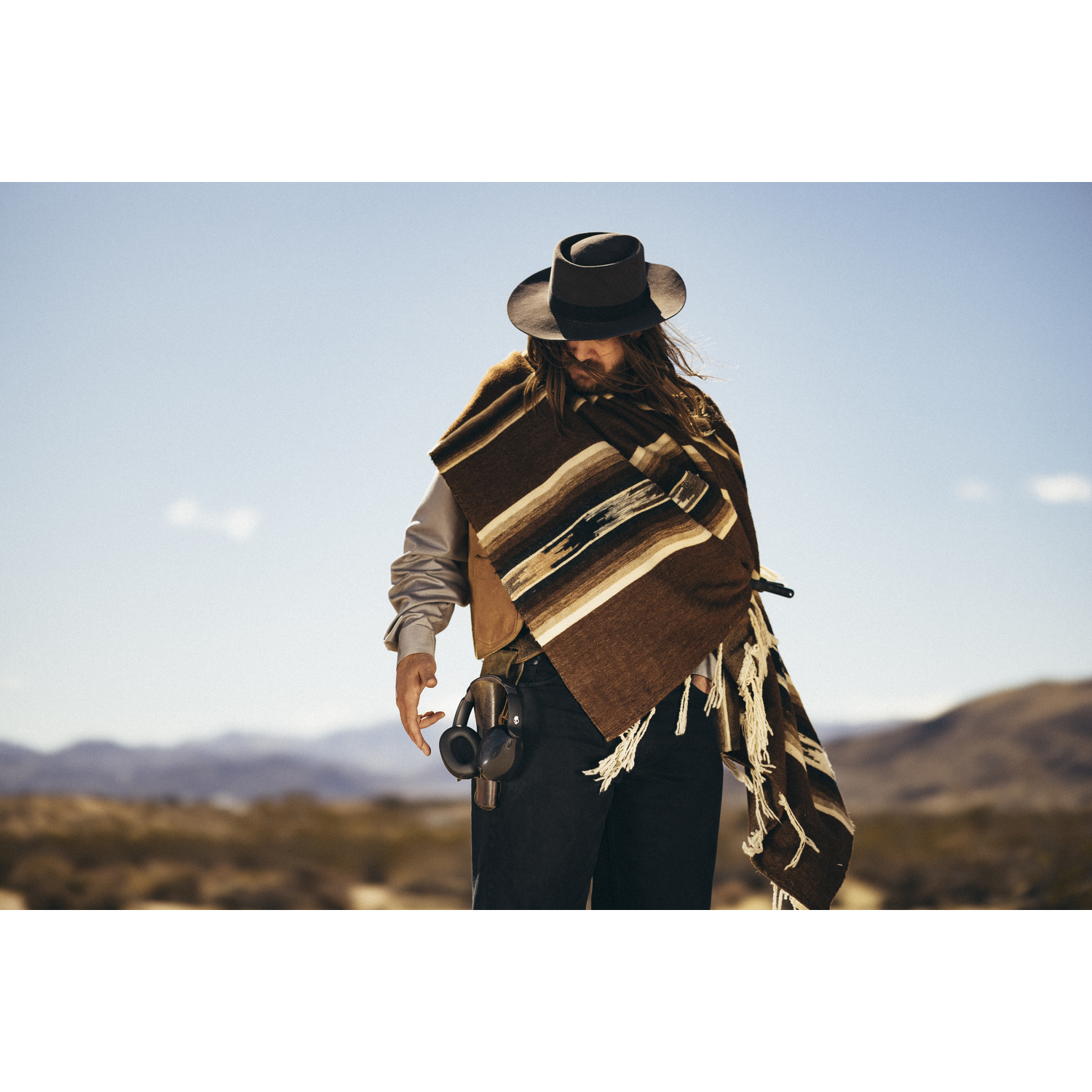 Un homme en chapeau et poncho se tient dehors. Il porte un pantalon noir et un holster. Le fond est un paysage désertique.
