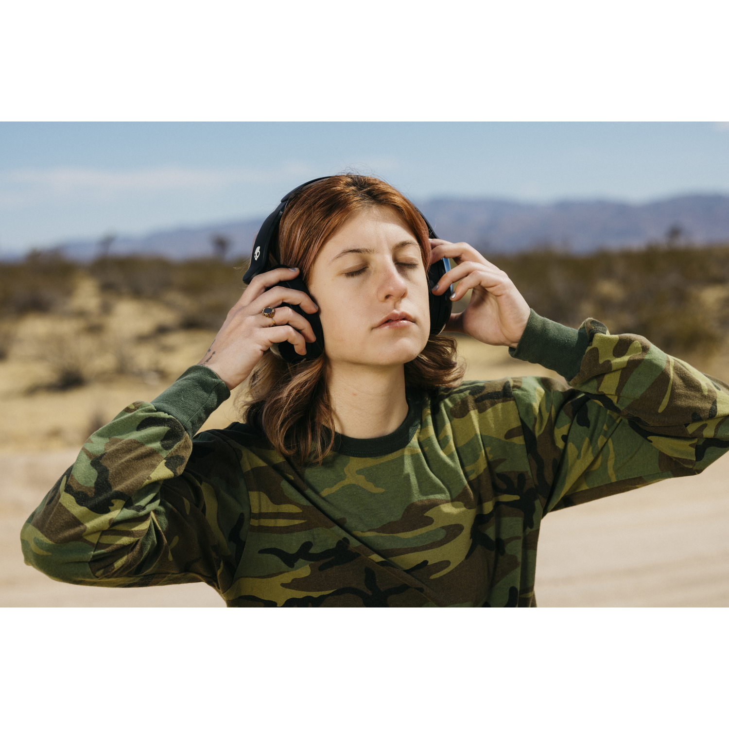 Une personne aux yeux fermés portant un casque et une chemise camouflage dans un cadre désertique.